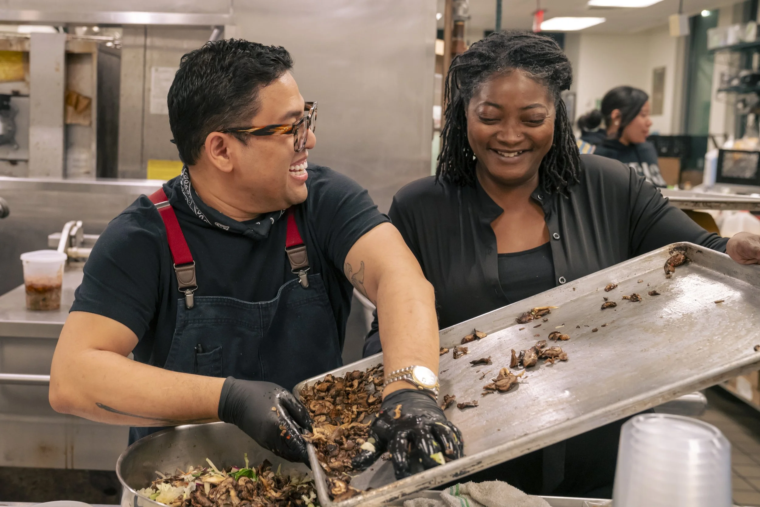 Two people working together in a kitchen, preparing food on a large metal tray, with one person wearing black gloves and the other smiling. There is a woman in the background preparing food as well.