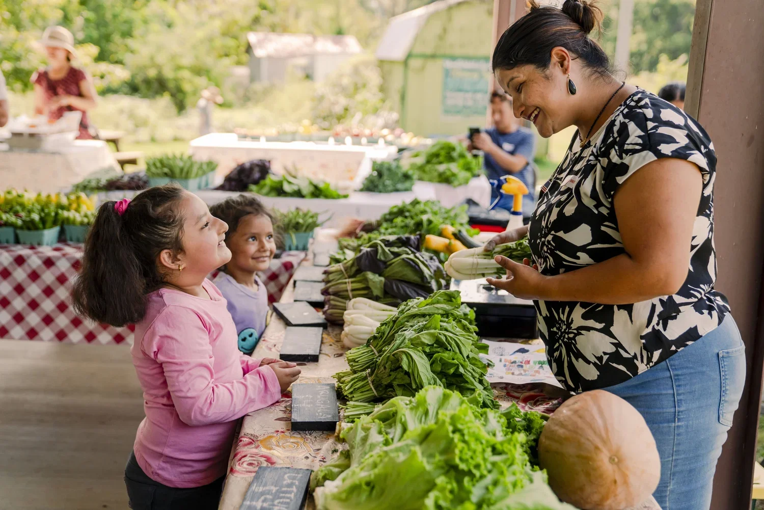 A woman selling fresh vegetables at a farmers market stall is interacting with two young girls who are smiling and looking at her.