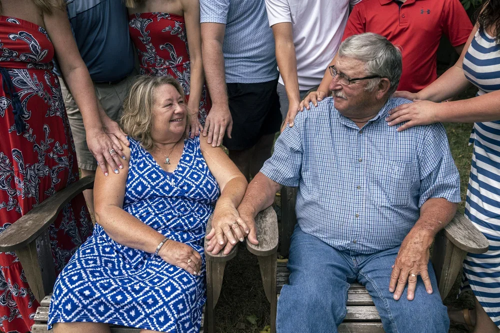 An elderly couple sitting on wooden chairs outdoors, holding hands and smiling at each other, surrounded by their family members.
