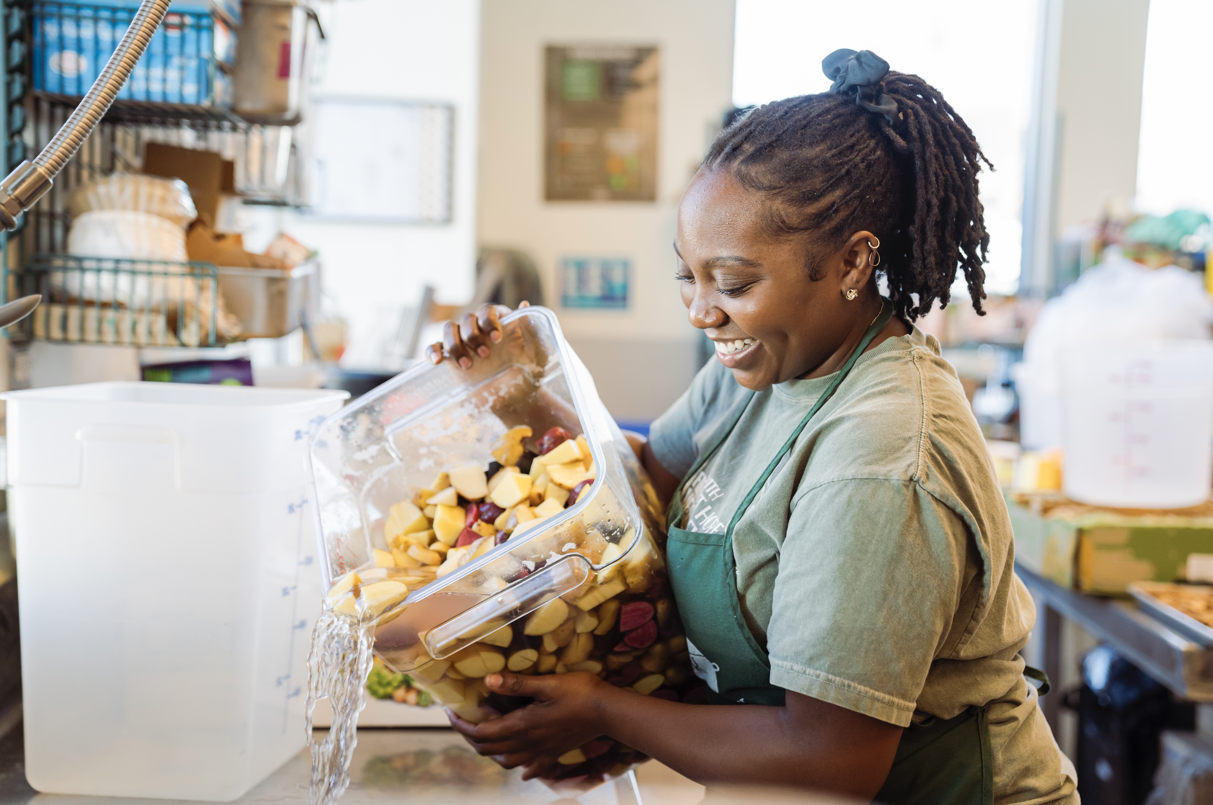 A woman with dreadlocks wearing earrings and a green apron smiling while pouring water from a container filled with cut fruits into a sink in a kitchen or cafeteria setting.