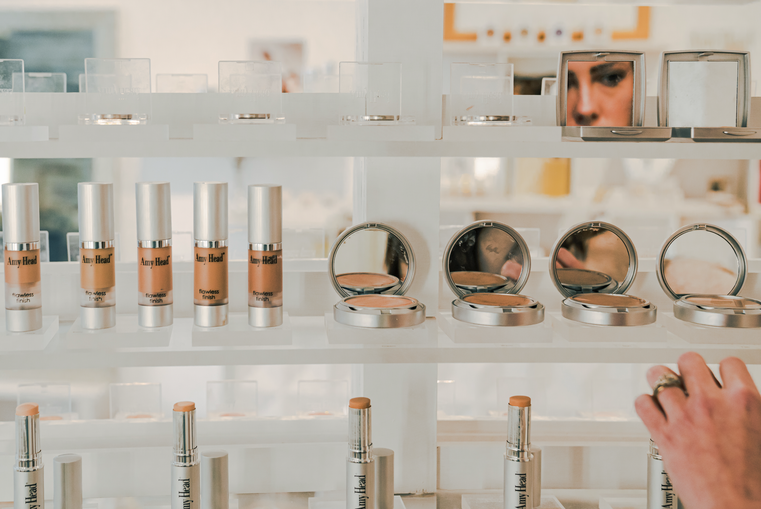 Display of various Amy Head cosmetics including foundation bottles and compact powders arranged on clear shelves.