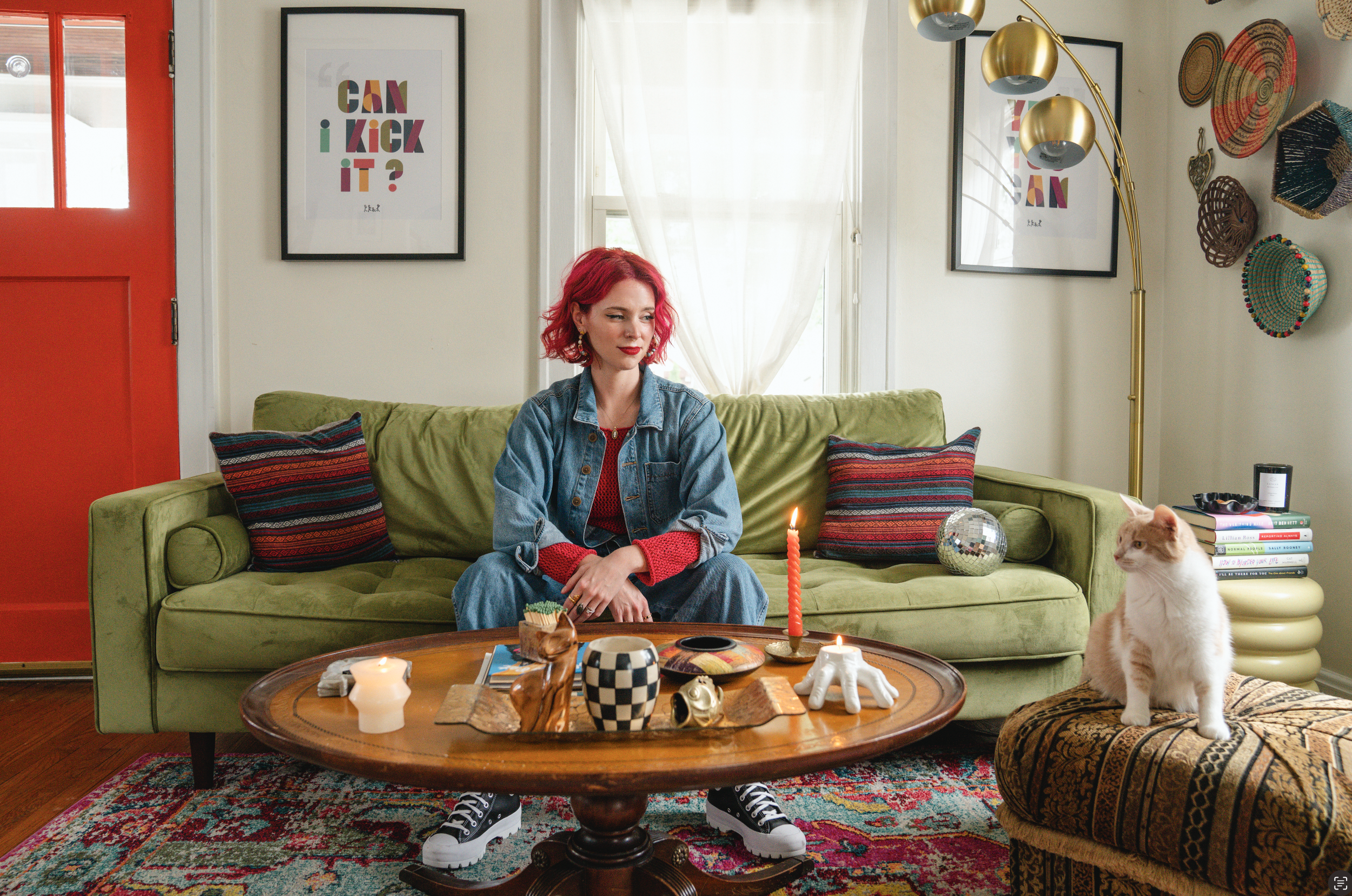A woman with red hair sits on a green couch in a living room decorated with framed artwork, woven baskets, and a floor lamp with gold shades. She is wearing a denim jacket over a red sweater. In front of her is a wooden coffee table with various deco