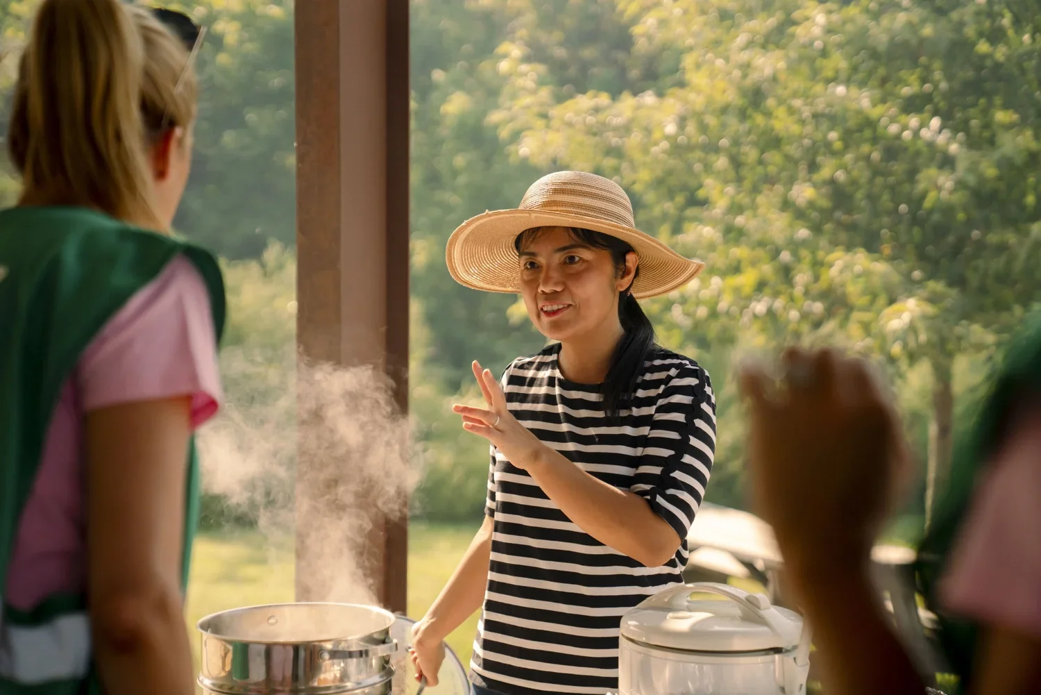 A woman wearing a large straw hat and a black and white striped shirt talking to other people at an outdoor gathering, with steam rising from a food container nearby, in a green, wooded area.