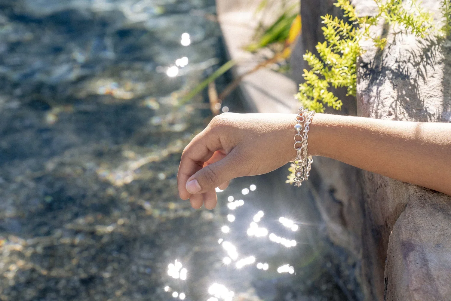 A person's hand extended over a water feature with sunlight reflecting off the surface, wearing silver bracelets, and surrounded by green plants and a rocky edge.