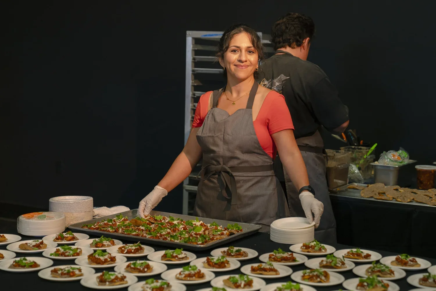 A woman smiling at a food event, placing food on small plates with an array of prepared dishes in front of her.