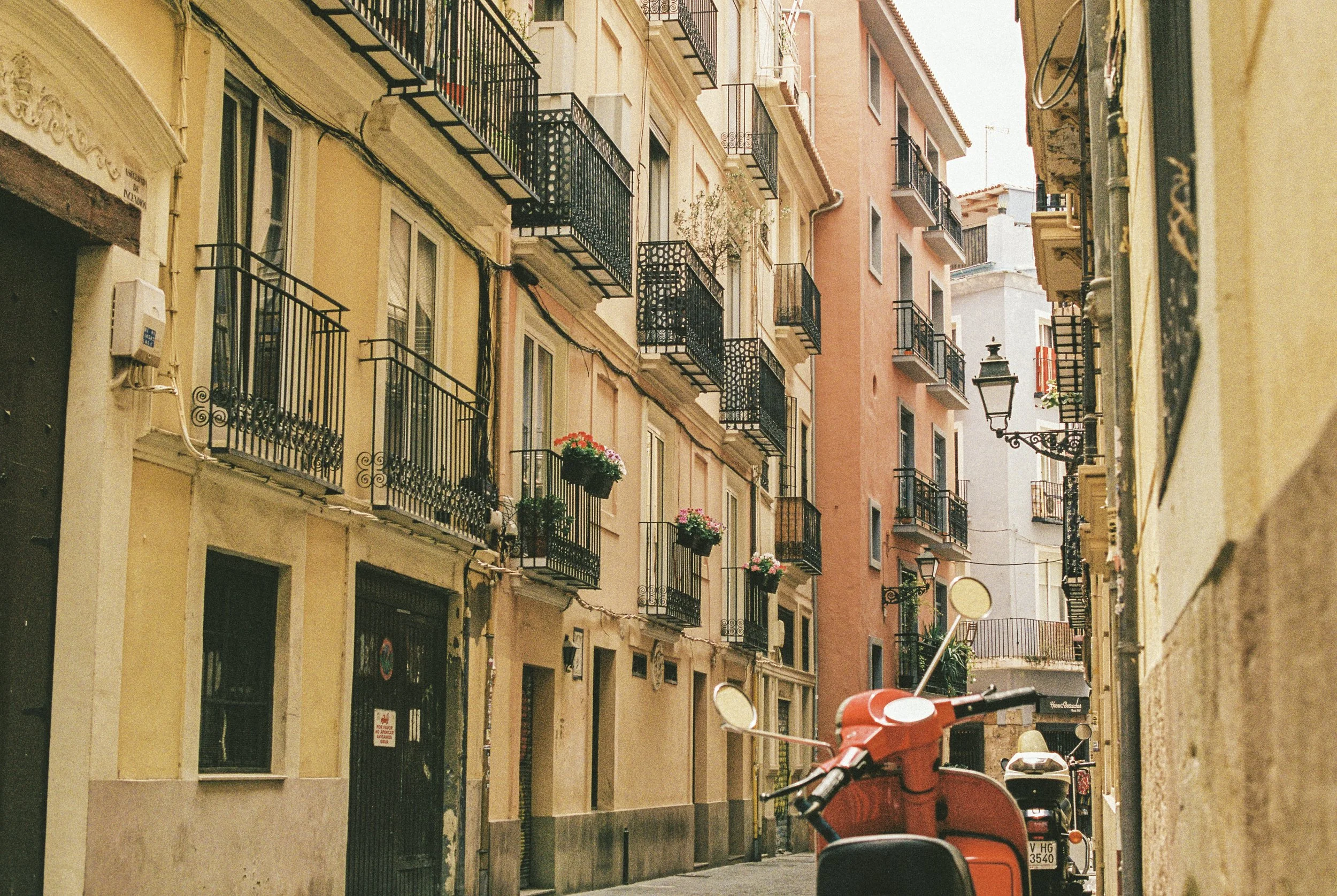 Narrow street lined with colorful buildings, each with black wrought iron balconies decorated with flower pots. A street lamp is visible, and a red scooter is parked on the sidewalk.