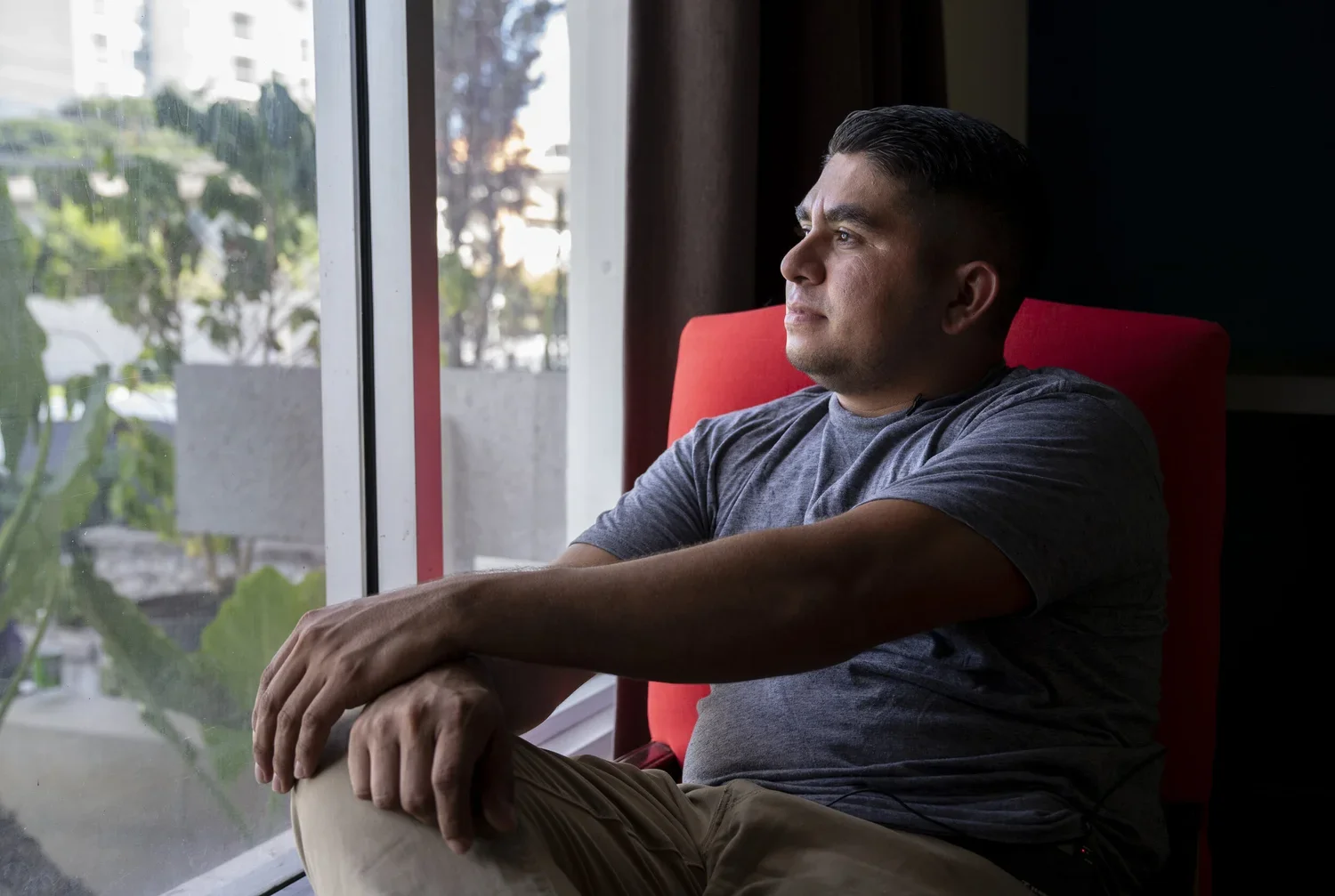 A man sitting in a chair near a window, looking outside with a contemplative expression.