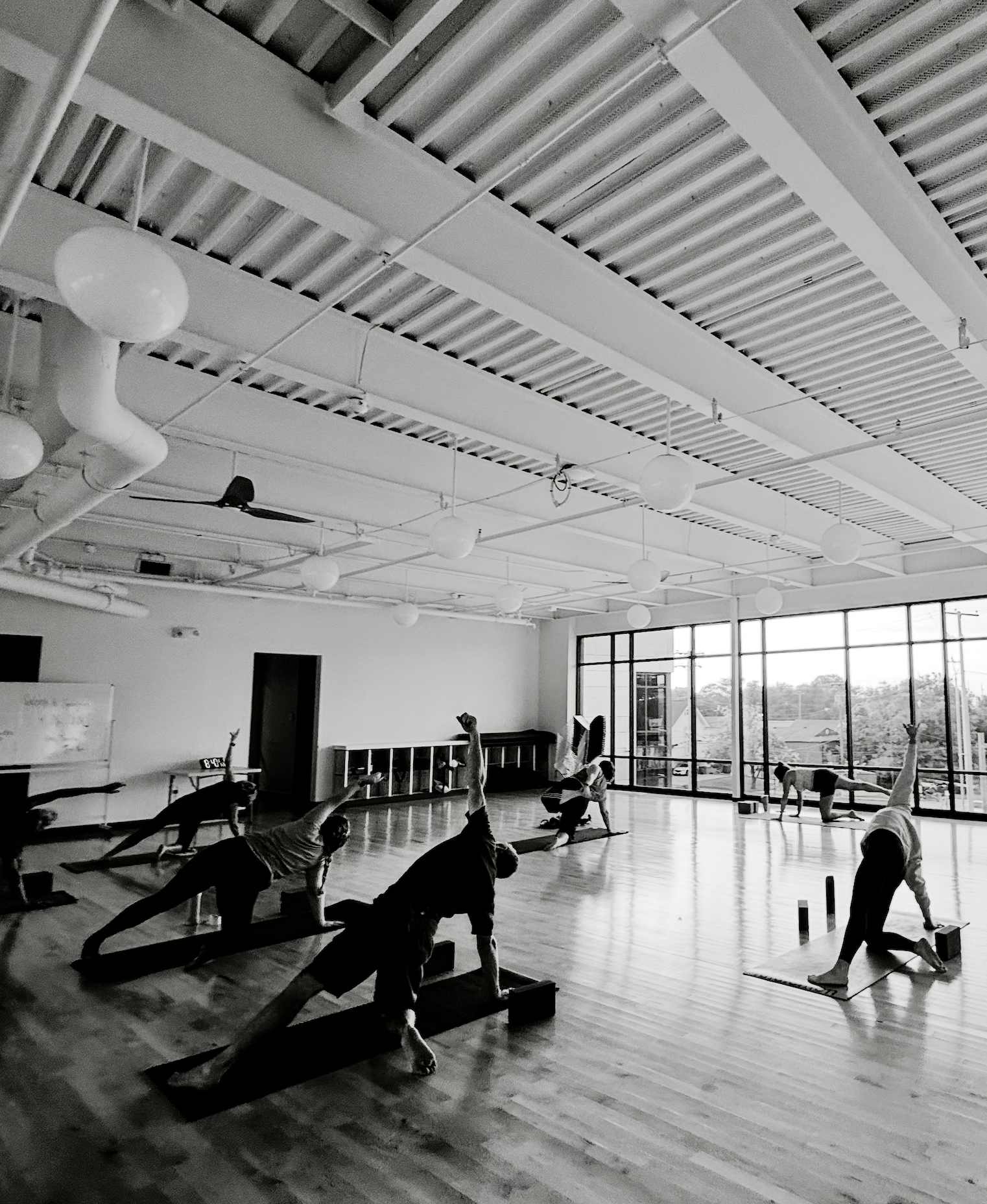 People practicing yoga in a spacious studio with large windows, wooden floors, and a high ceiling with exposed beams and hanging light fixtures.