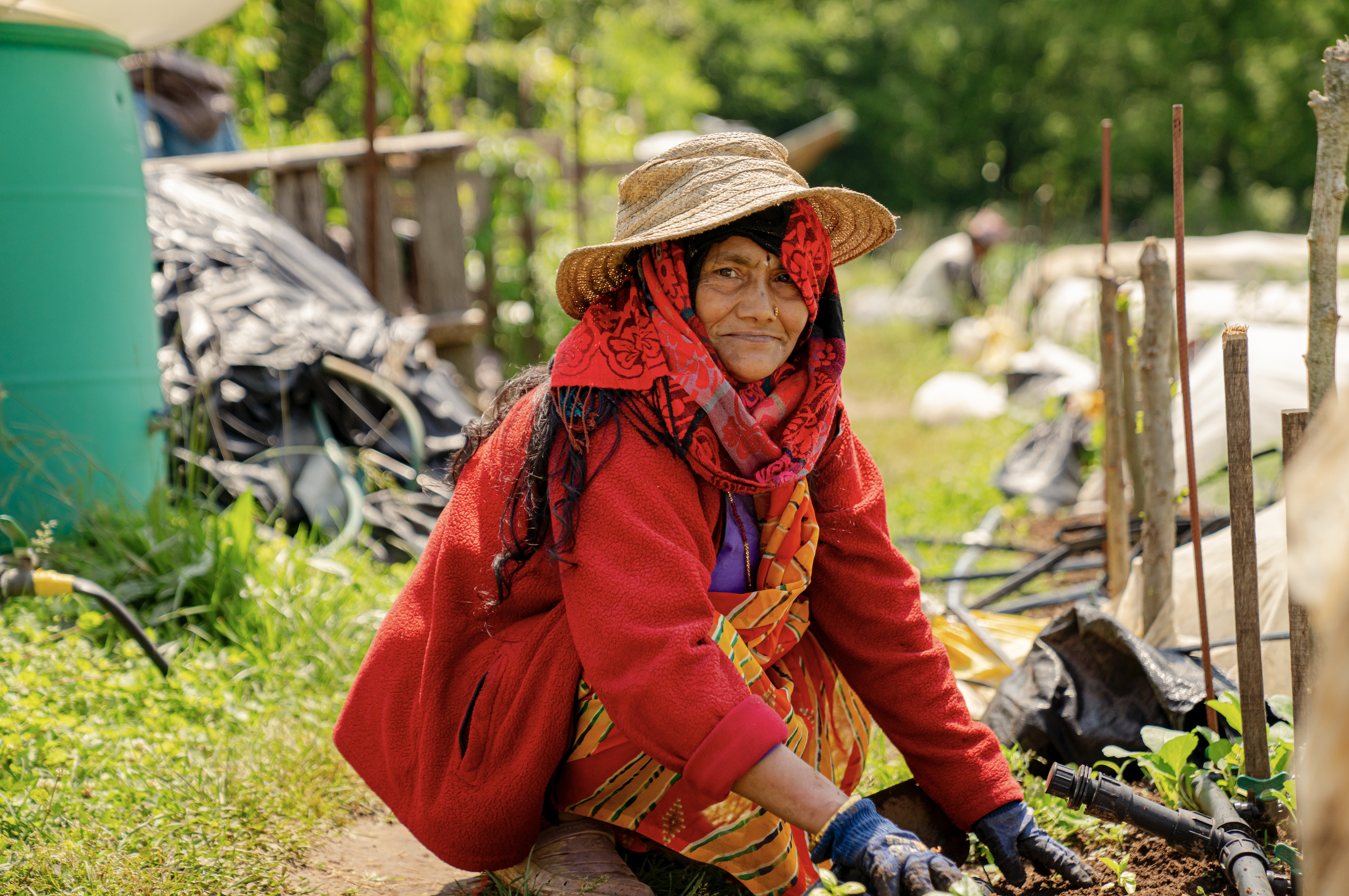 A woman wearing a straw hat, red scarf, red fleece, and colorful skirt planting or working in a garden, with greenery and gardening tools around her.