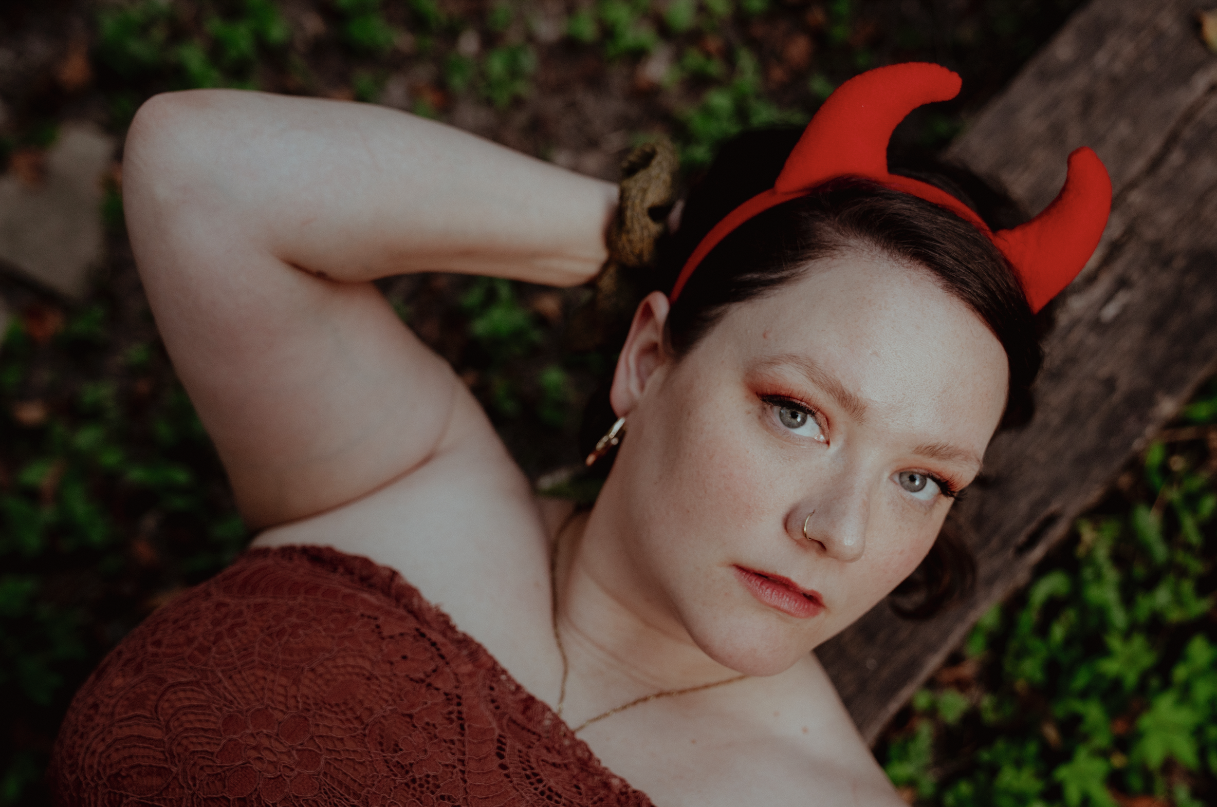A woman with fair skin and blue eyes wearing a red devil horns headband and a lacy red top, lying on the ground near a tree trunk with green foliage in the background.