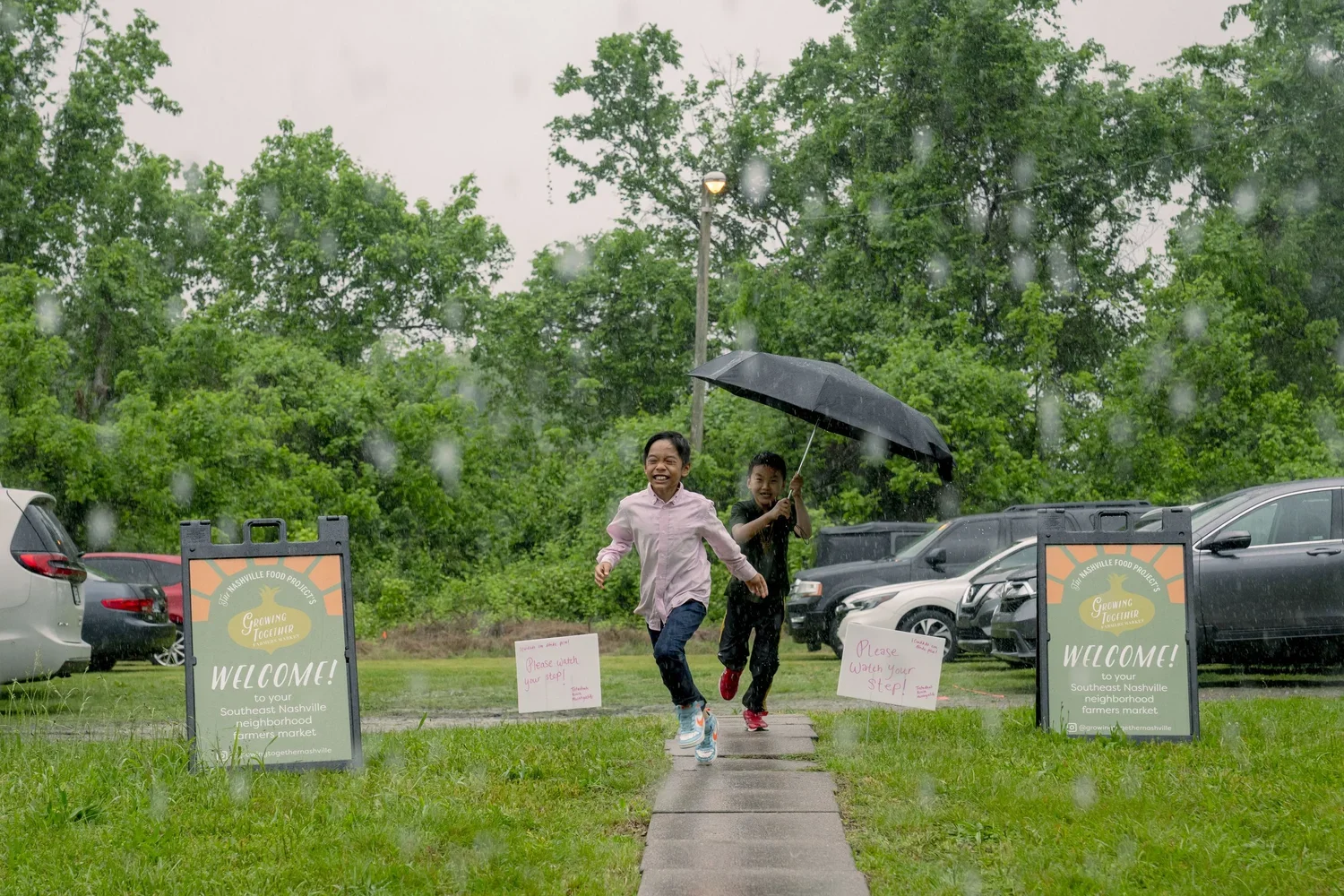 Two children running on a rain-soaked sidewalk, one holding an umbrella, with cars parked on a grassy area and green trees in the background.