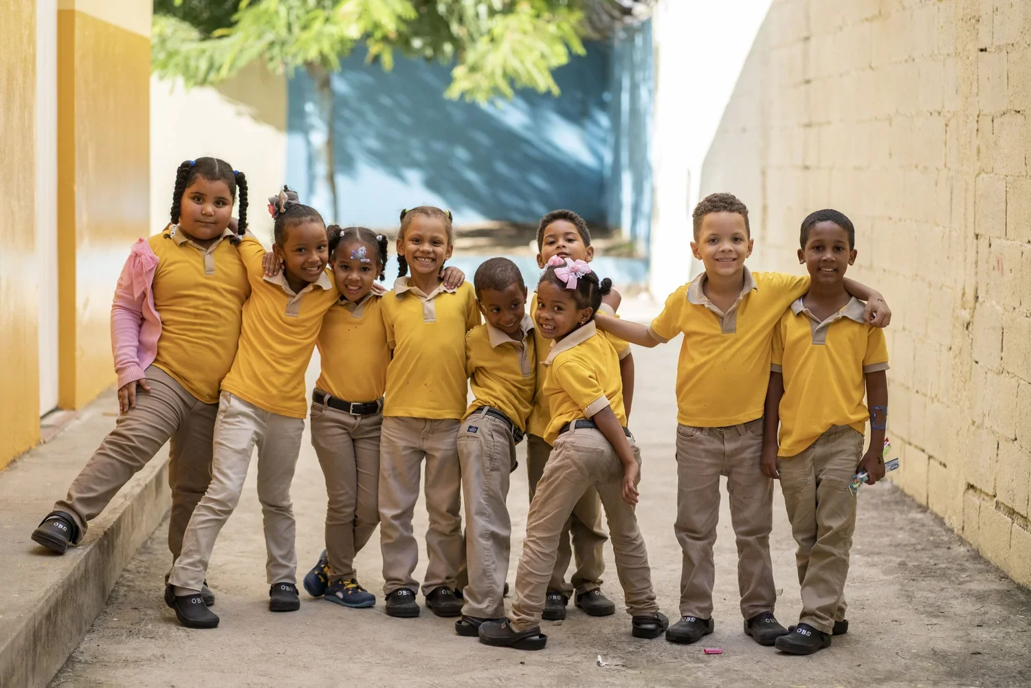 Group of ten diverse children in yellow school uniforms standing arm-in-arm in an outdoor corridor, smiling at the camera.