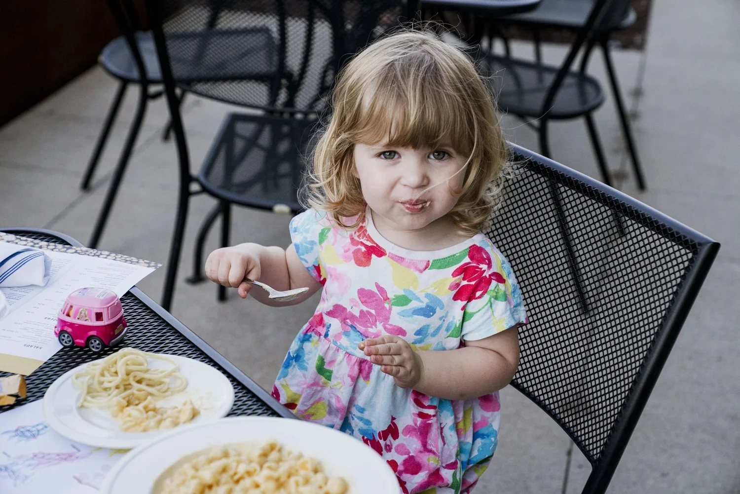 A young girl with curly blonde hair wearing a colorful floral dress sitting at an outdoor table, holding a spoon and looking at the camera with a playful expression. The table has plates of pasta, a pink toy car, and a paper menu.