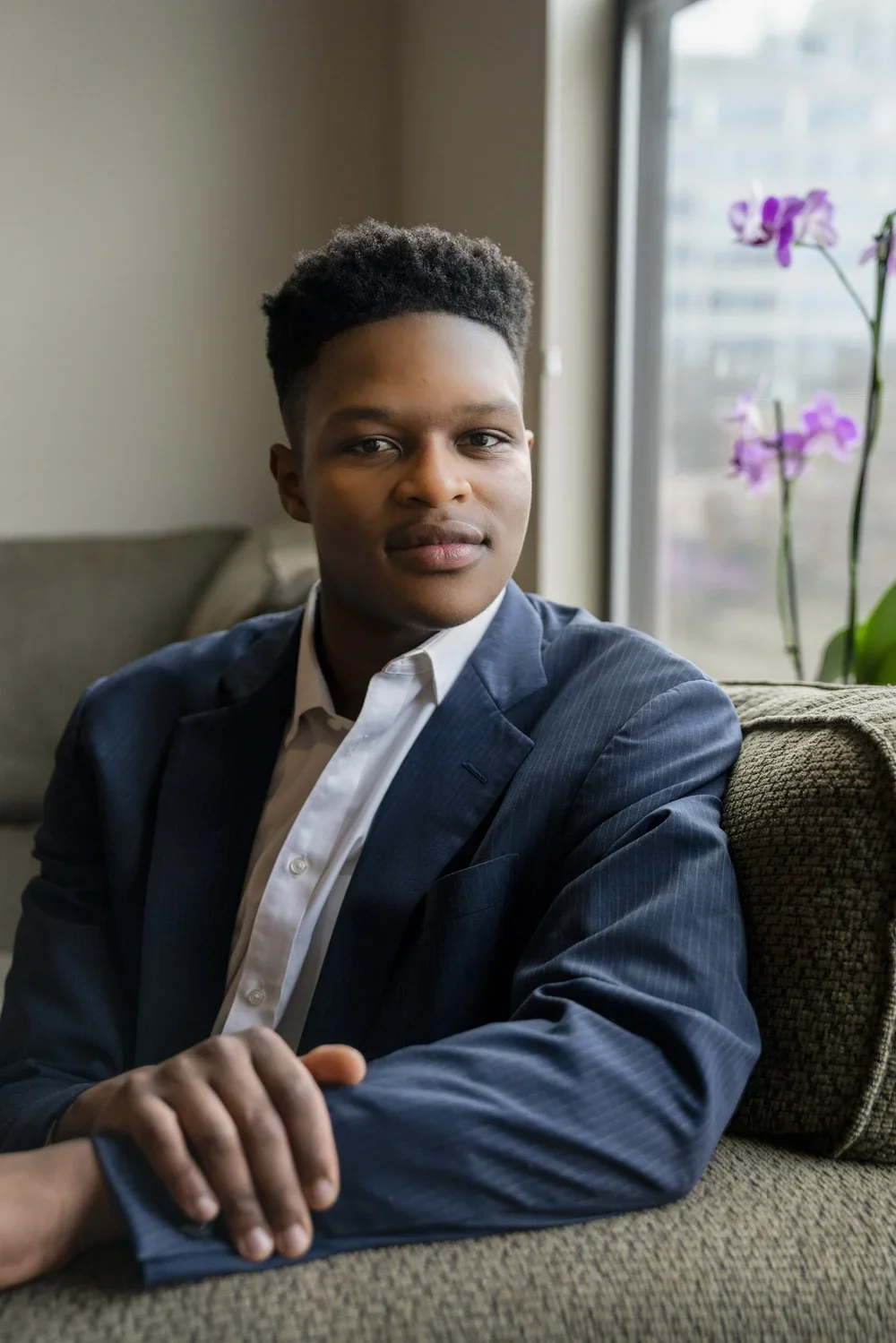 A young man with dark skin and short curly hair, wearing a formal dark blue suit and white shirt, sitting on a sofa in a room near a window with purple flowers in the background.