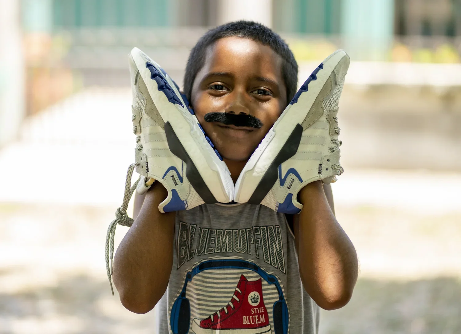 A young boy with a fake mustache on his face holding a pair of sneakers on his shoulders, smiling at the camera.