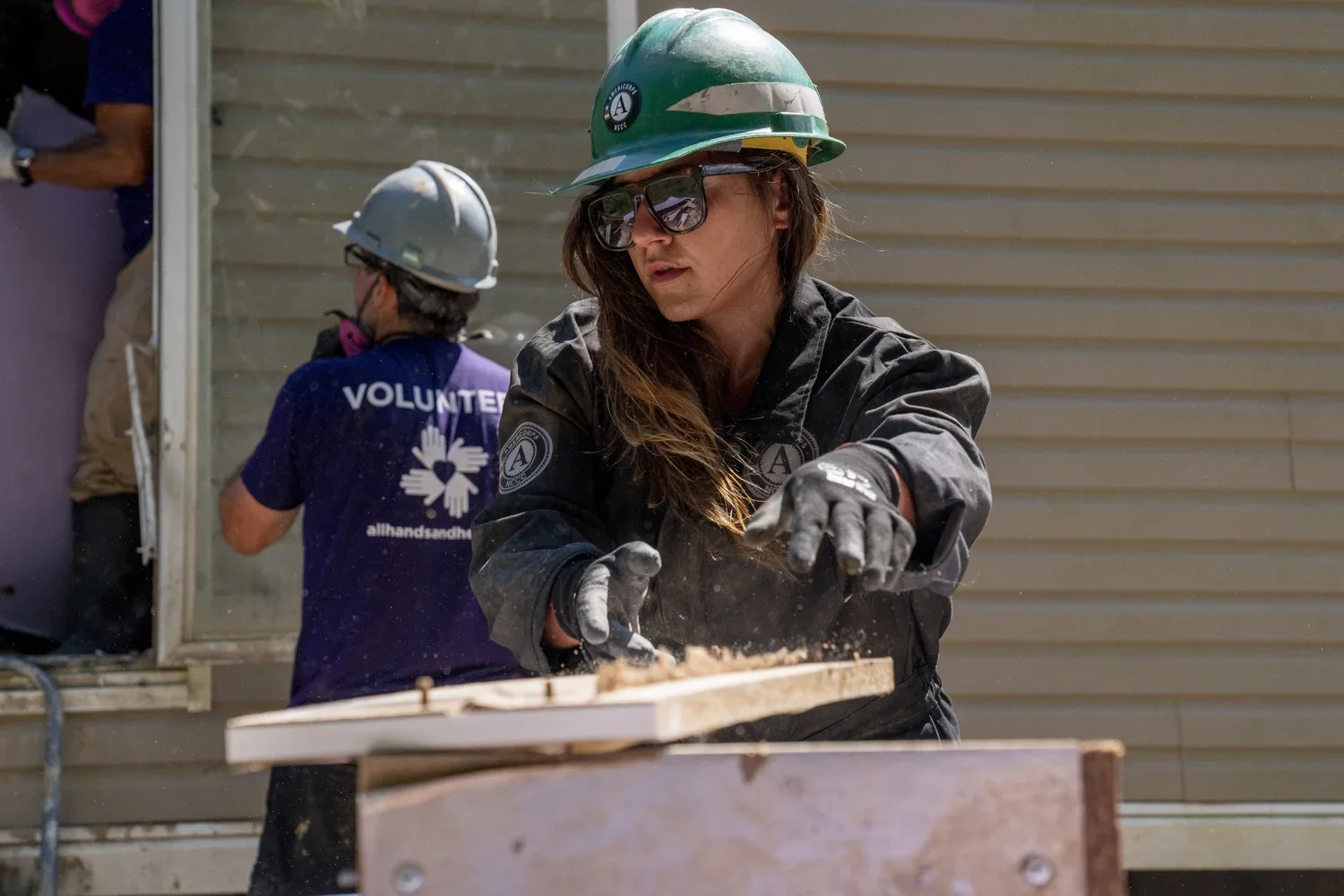 A woman wearing a green hard hat, sunglasses, and black work gloves is working on a woodworking project outdoors. Behind her, another volunteer is also wearing a helmet and a purple shirt with 'VOLUNTEER' written on the back, indicating they are help