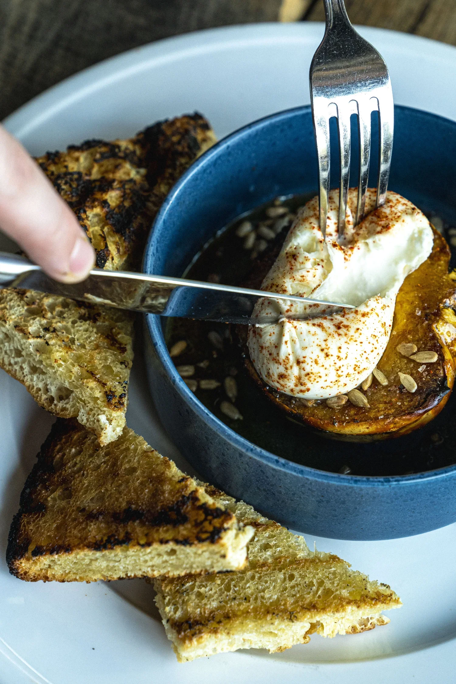 Slices of toasted bread with a golden-brown crust and a soft, airy interior, served with a bowl of hummus topped with a dollop of whipped topping or yogurt, sprinkled with paprika. A fork is inserted into the hummus, and a hand is holding a piece of 