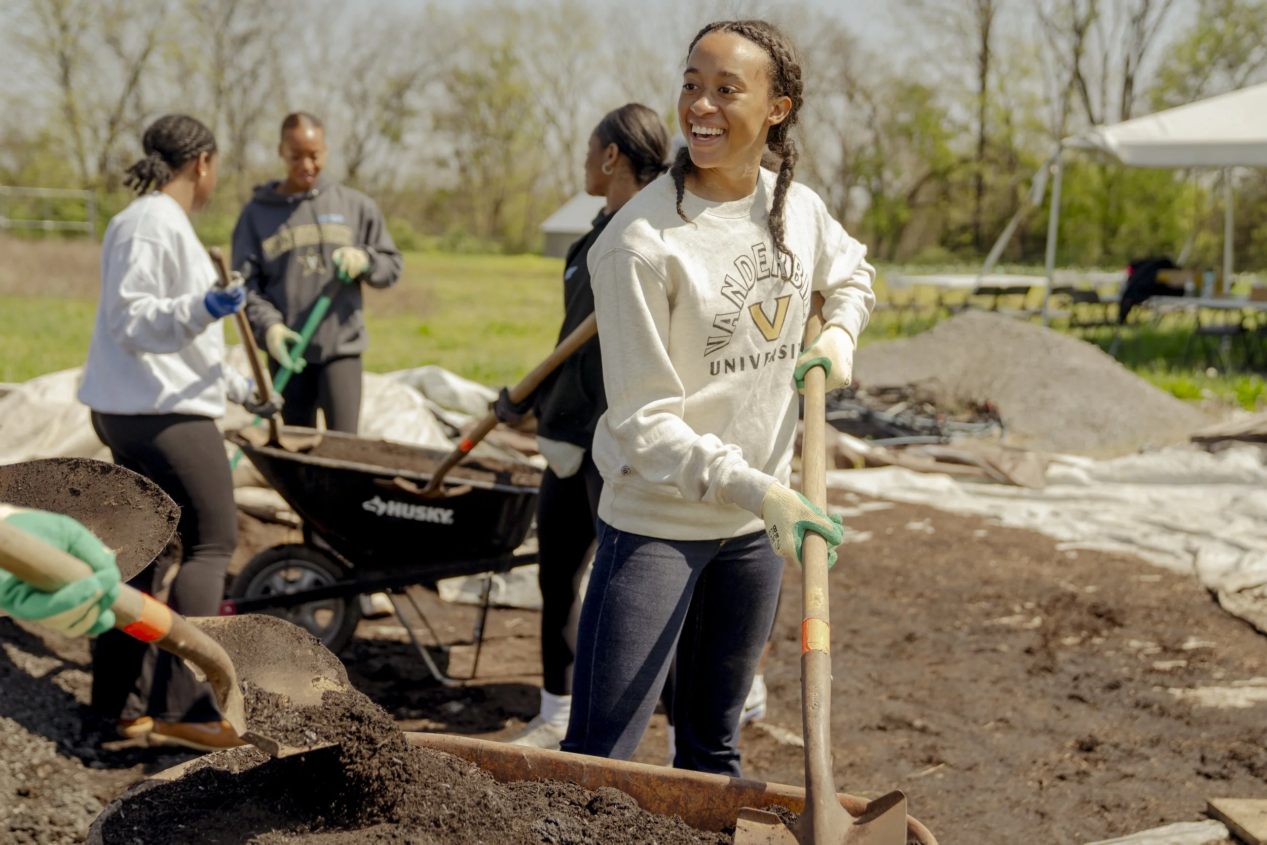 Young woman with braids smiling while shoveling dirt during a community outdoor project, with other volunteers working in the background.
