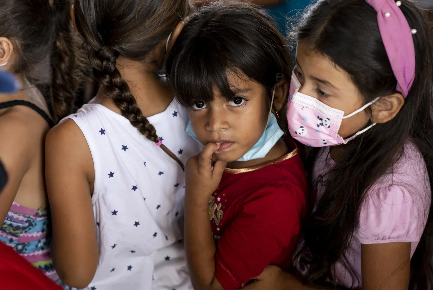 Group of young girls, some wearing face masks, closely gathered with one girl looking directly at the camera, finger to her lips, in an indoor setting.
