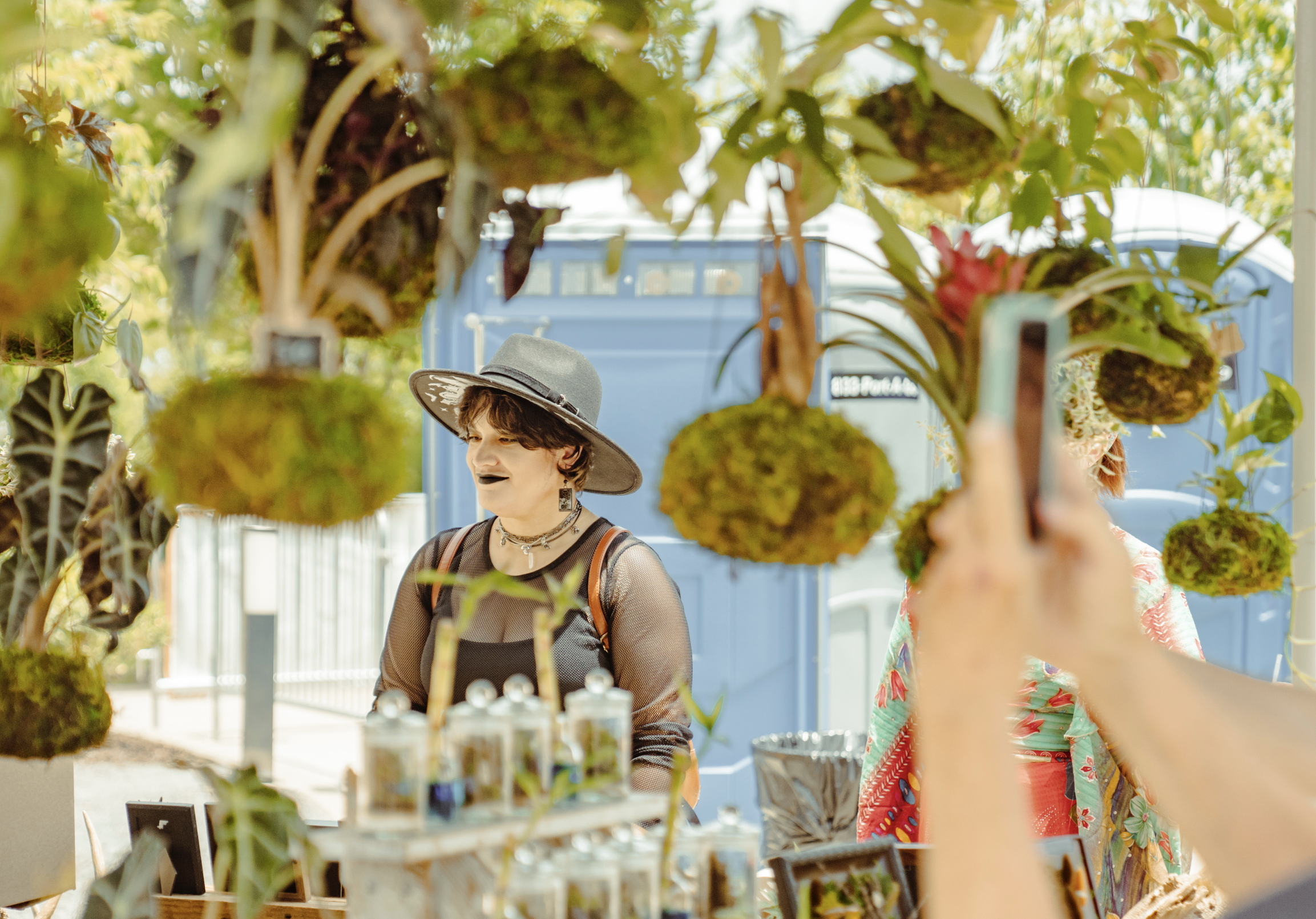 Person wearing a wide-brimmed hat with a band and earrings, shopping at an outdoor market with plants and art supplies, with a blue bus in the background.