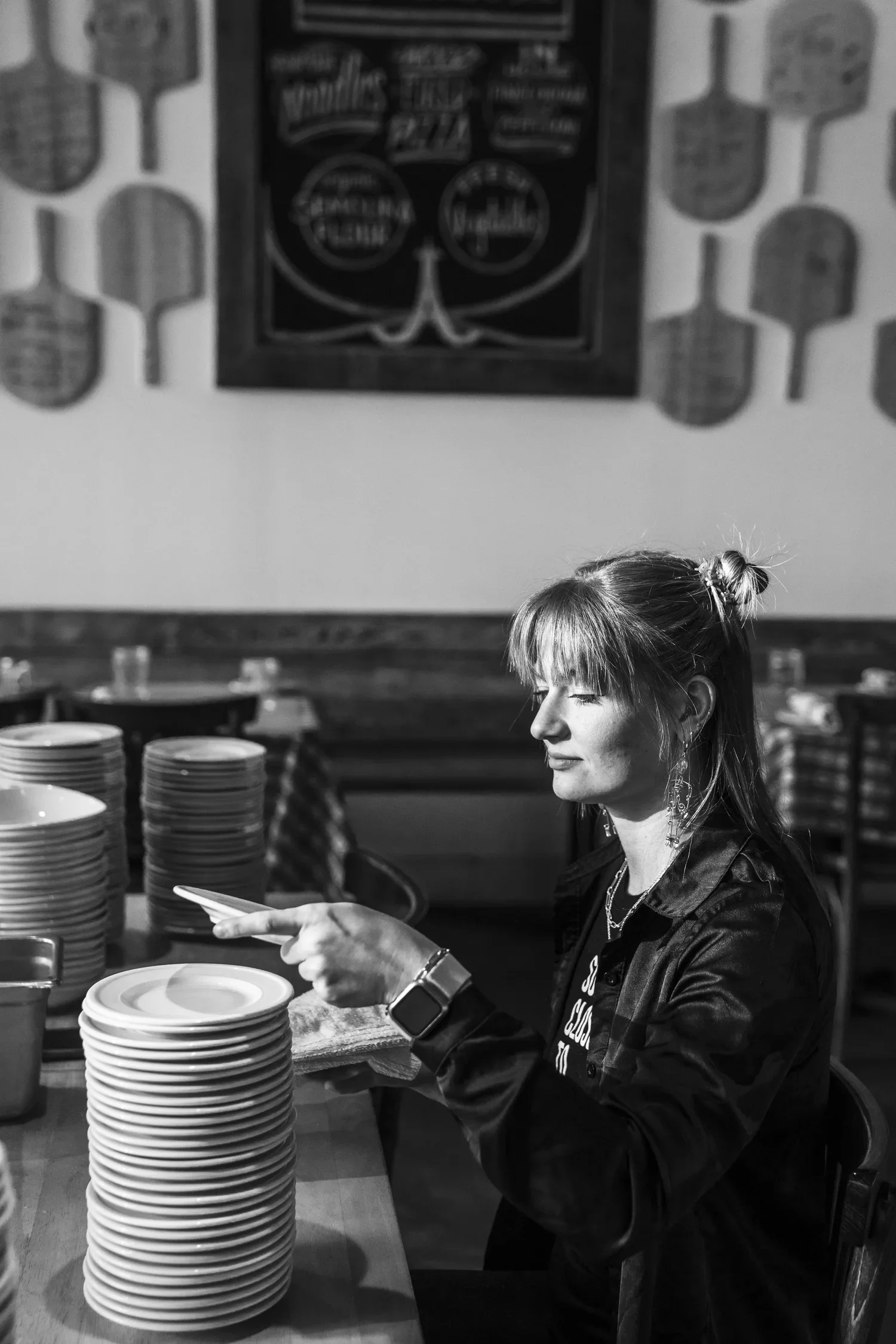 A woman with long hair styled in a top knot is sitting at a table in a restaurant or cafe, looking at her phone. She is wearing a black jacket and a smartwatch. There are stacks of plates and cups on the table. The background features a wall with a d