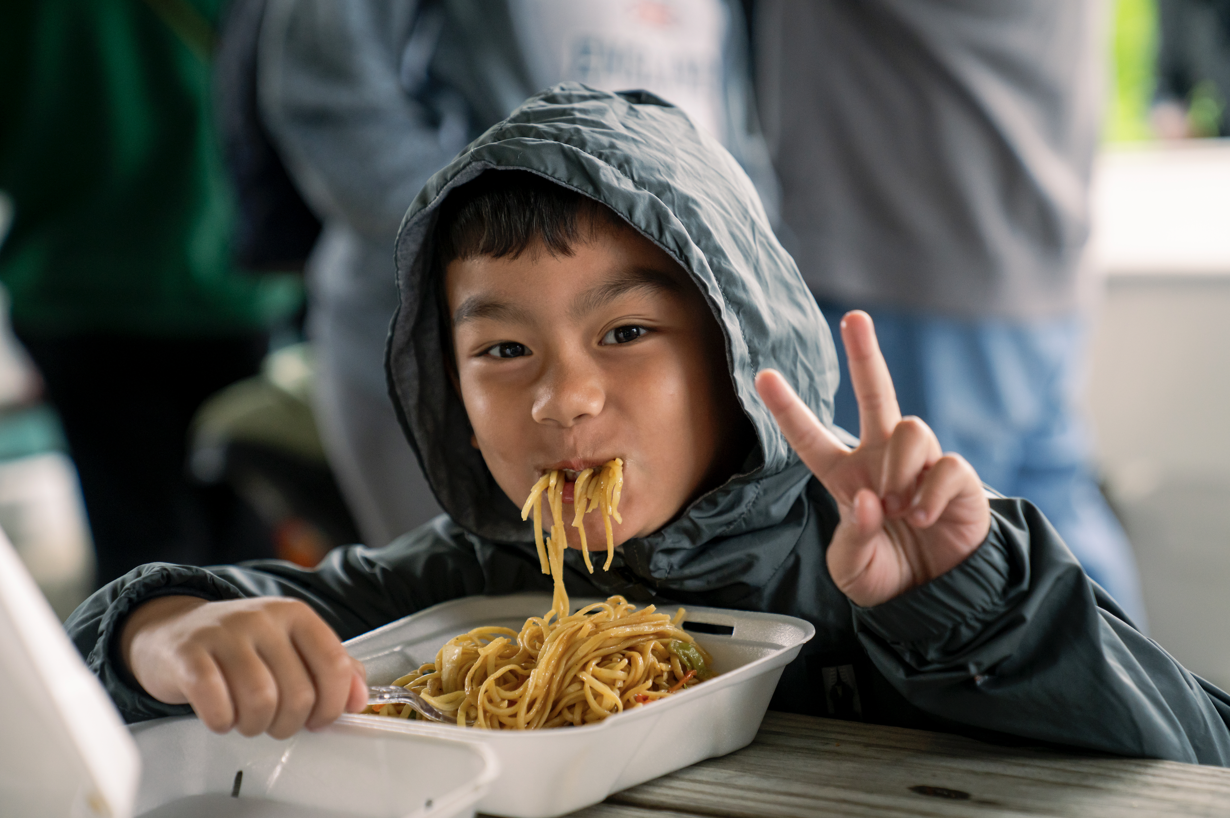 A young boy in a gray hooded jacket is eating spaghetti with vegetables from a white takeout container, holding a fork in his right hand and making a peace sign with his left hand, sitting at a table.
