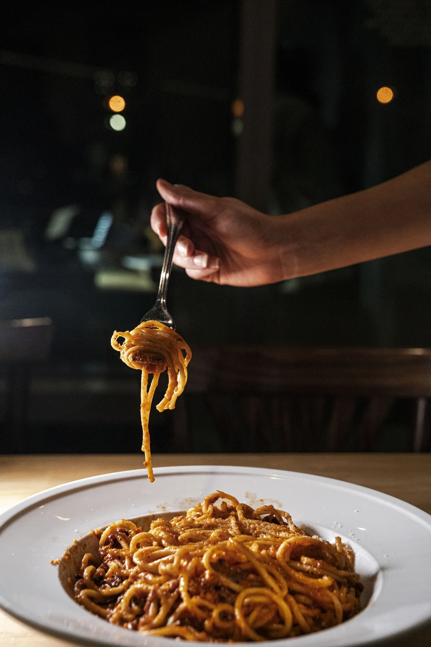 A hand holding a fork with a noodle strand above a bowl of spaghetti in a dark setting with blurred lights in the background.