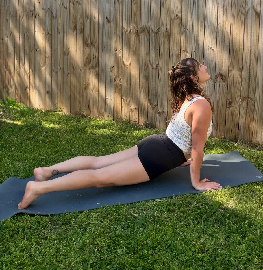 A woman practicing yoga outdoors on a black mat on the grass, performing Bhujangasana (cobra pose) with a wooden fence in the background.