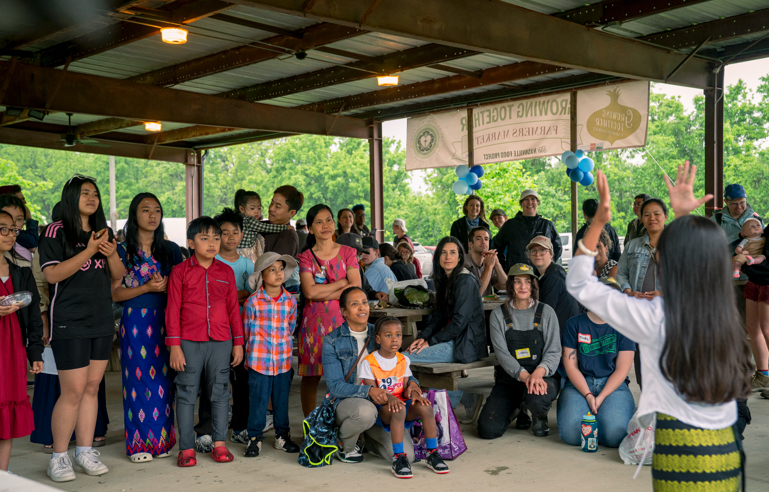 People gathered at an outdoor event, some standing and others sitting, with a banner and balloons in the background, under a metal roof with trees visible outside.