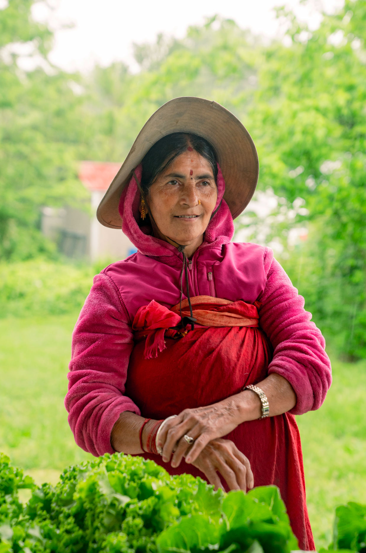 An elderly woman wearing a wide-brimmed hat and a pink hoodie over a red skirt, standing outdoors in a green landscaped area with lettuce in the foreground.