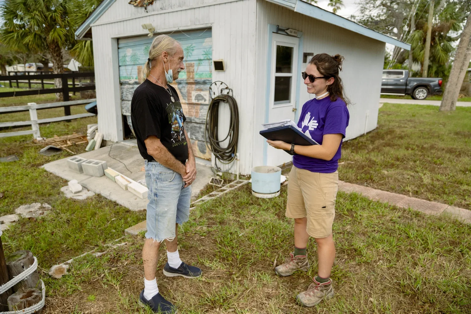 A young woman with glasses, wearing a purple t-shirt and beige shorts, holding a clipboard, talking to an older man with long white hair, wearing a black t-shirt, ripped shorts, and a face mask hanging below his chin, outside near a small white shed 