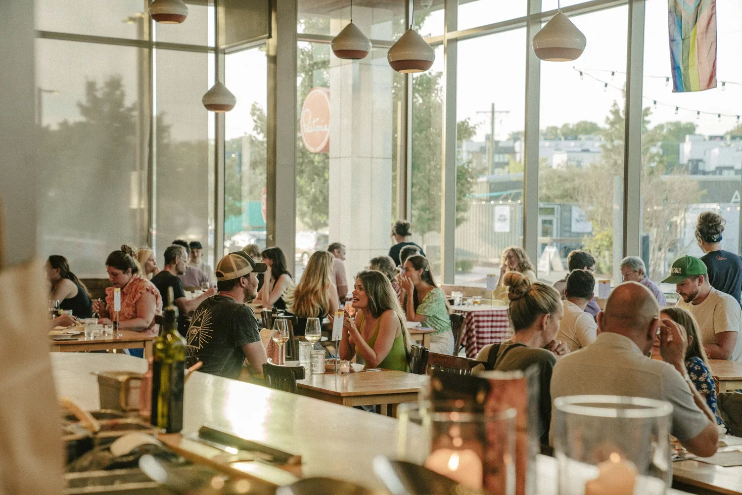 Crowded restaurant with people dining at tables near large glass windows, decorated with hanging pendant lights and a rainbow flag.