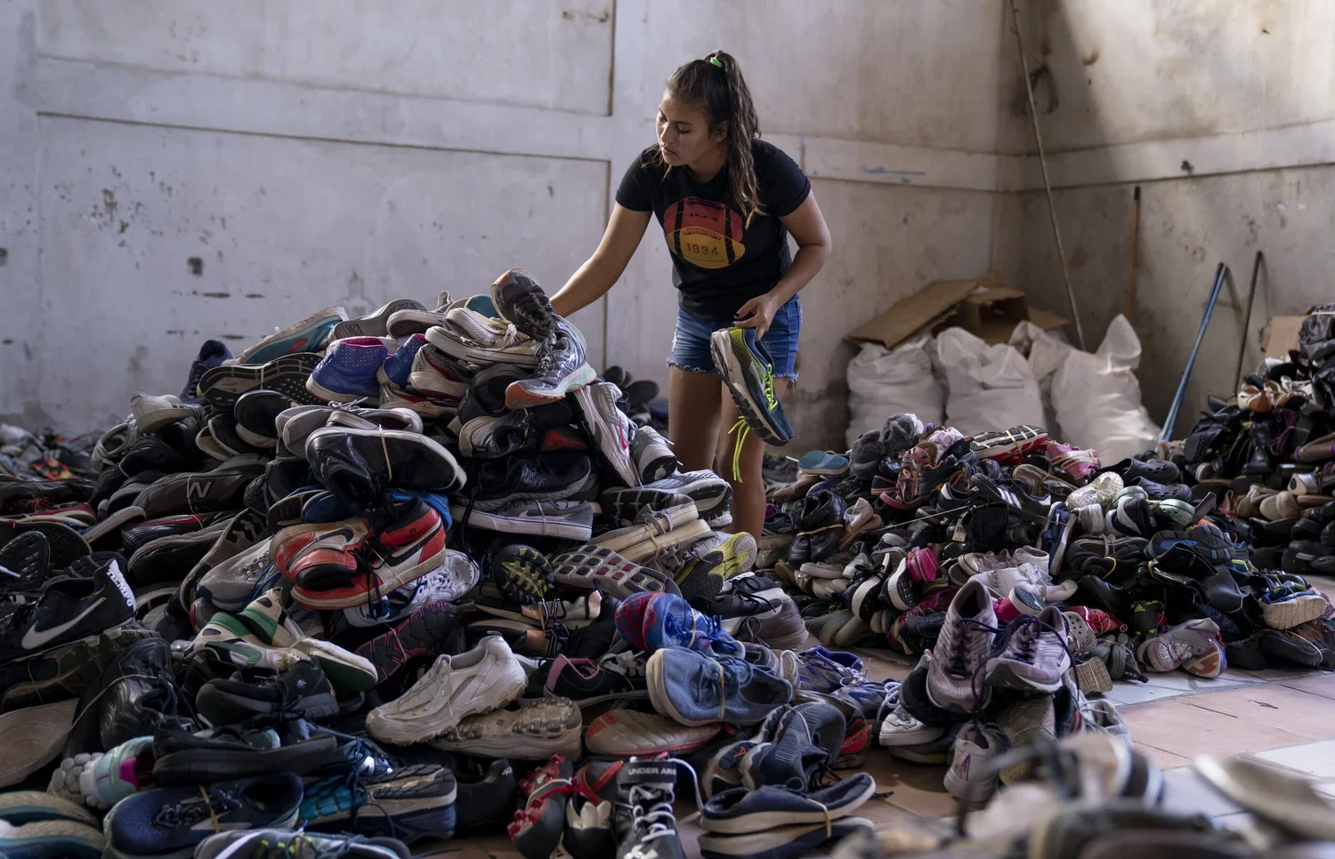 A young woman sorting shoe piles in a warehouse filled with scattered sneakers and shoe boxes.