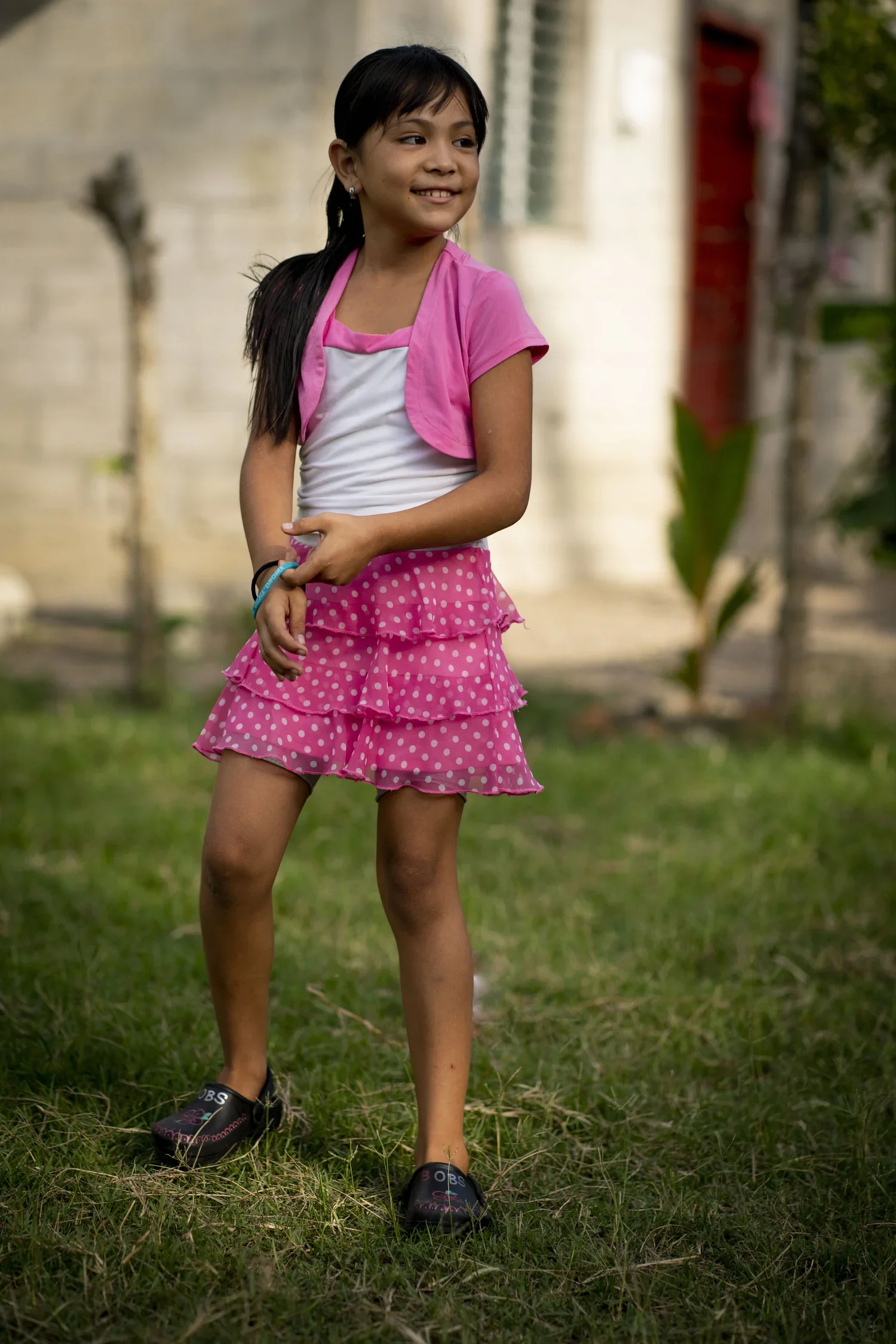 Young girl with long dark hair, wearing a pink and white top, pink polka dot ruffled skirt, black shoes, standing outdoors on grass with a blurred background of trees and buildings.