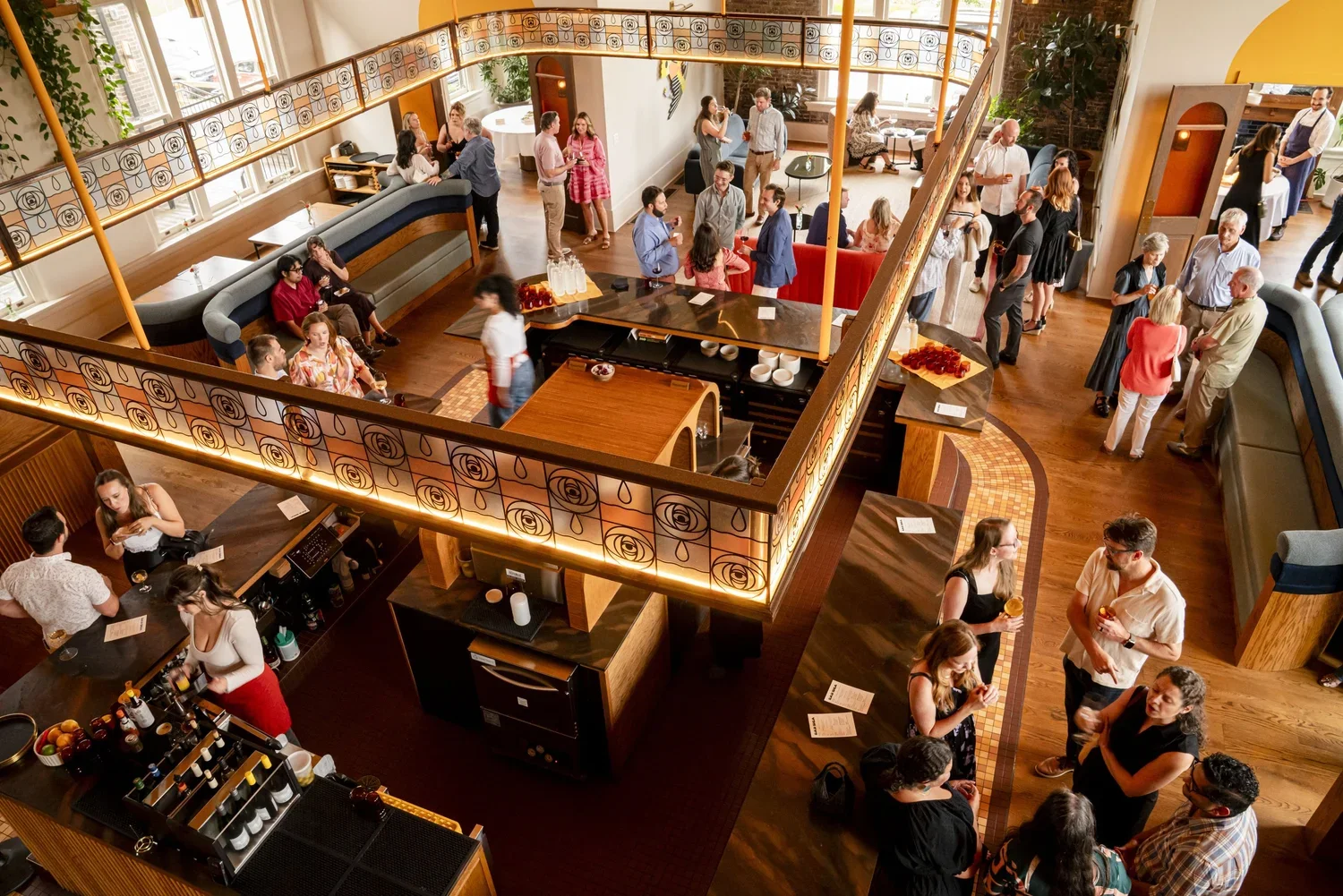 People socializing and enjoying drinks in a warmly lit, multi-level restaurant or bar with wood floors and modern decor.