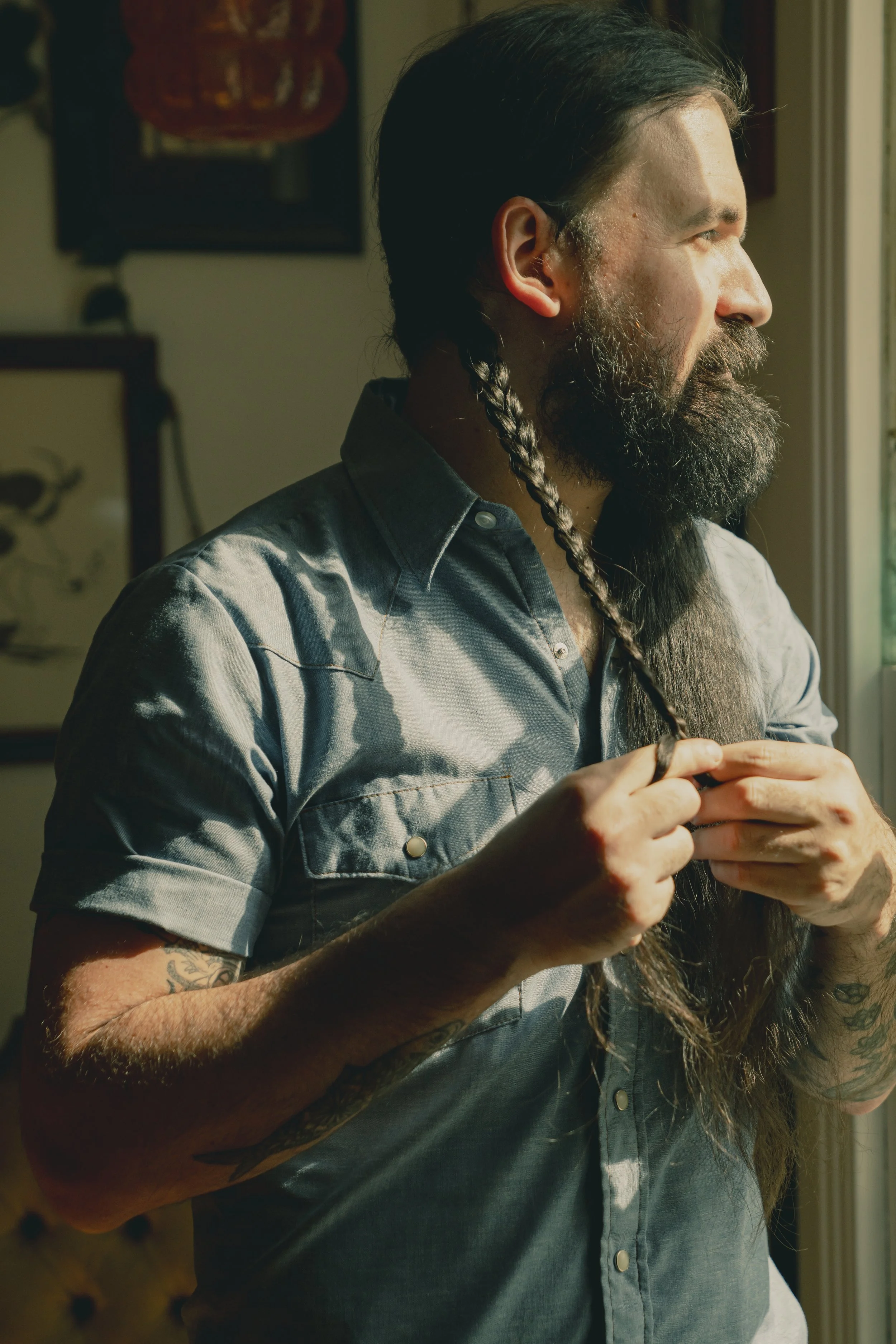 A man with a beard and long hair with a braid, wearing a denim shirt, is looking outside a window, illuminated by natural light.