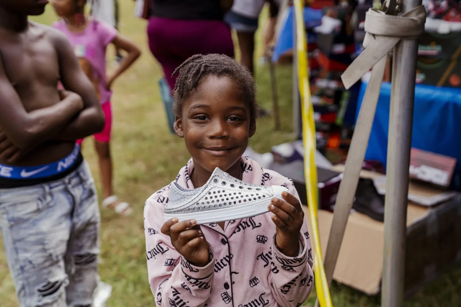A young girl smiling and holding a shiny silver sneaker in a crowded outdoor setting, with other children and market stalls visible in the background.