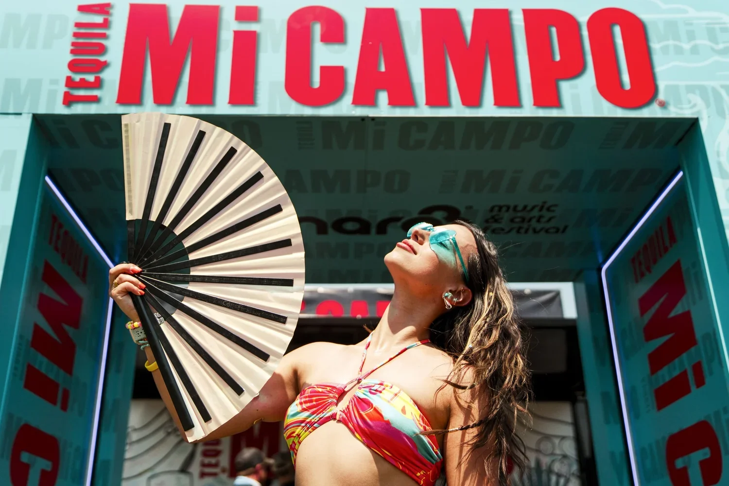 Woman in colorful swimsuit holding a fan, wearing sunglasses and earrings, standing under an archway with 'Mi Campo' signage at an outdoor event.