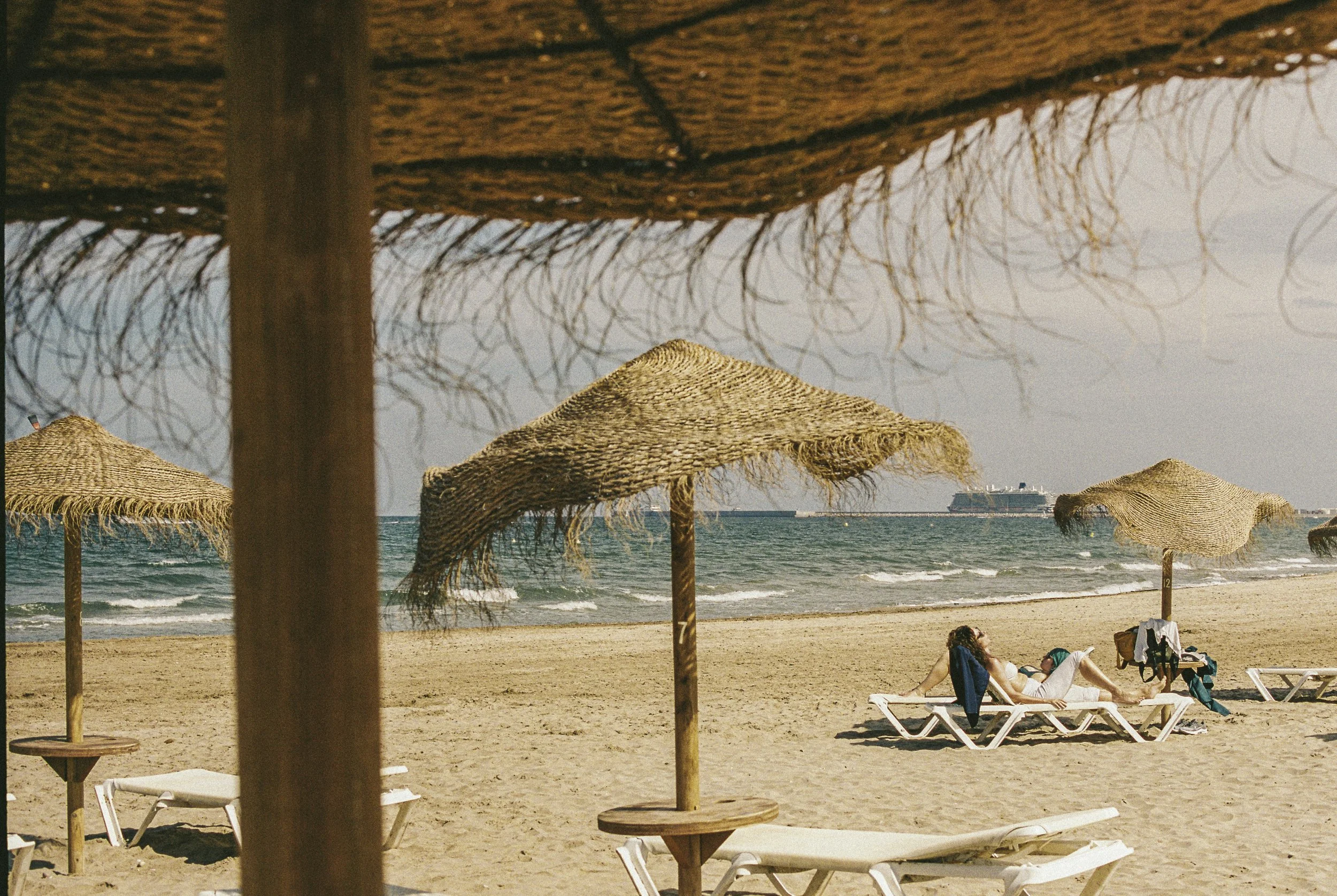 A beach scene with straw umbrellas and lounge chairs along the sandy shore, two people relaxing on chairs, with a large cruise ship in the distance on the water, seen under a sky with some clouds.