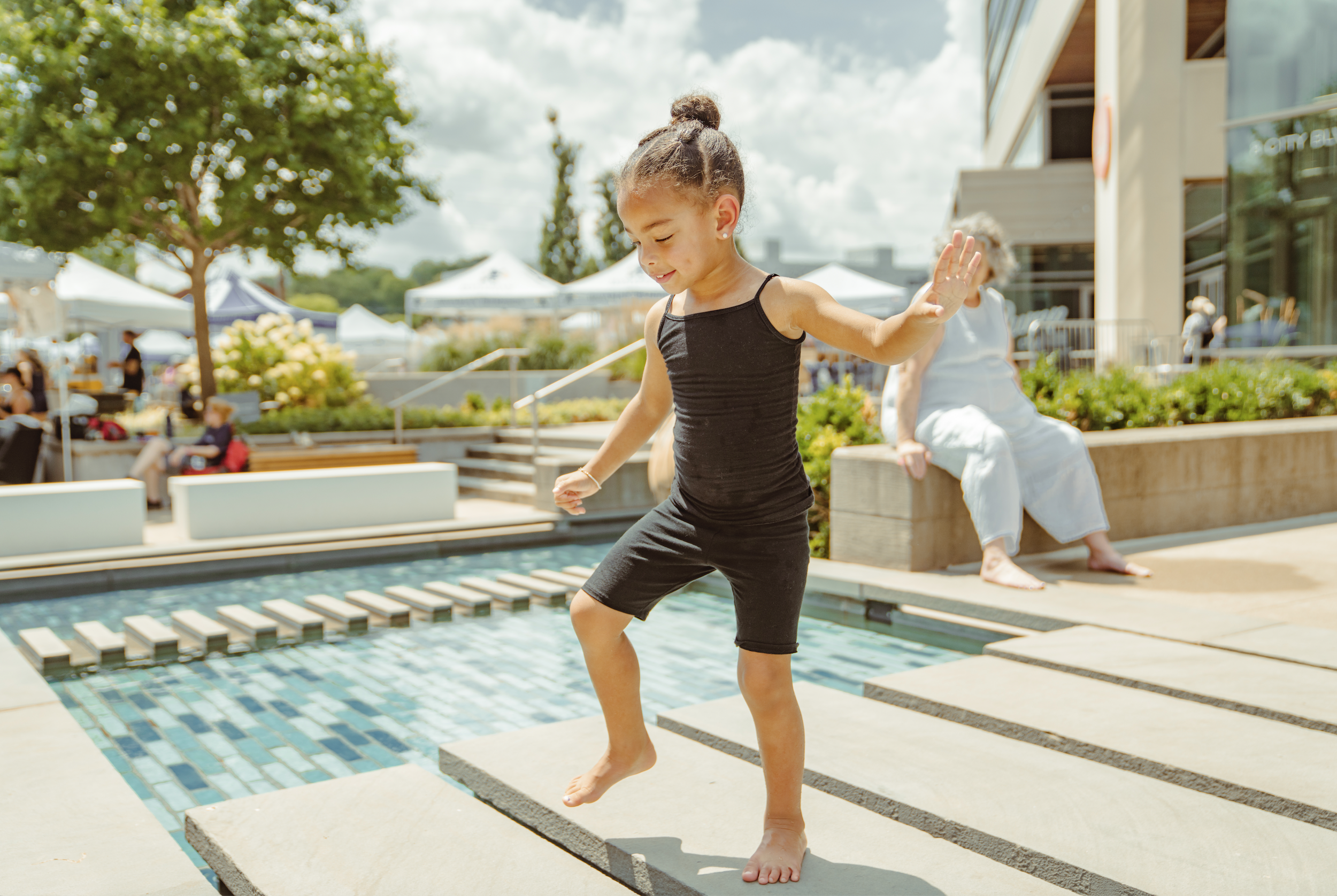 A young girl in a black dress balances on a floating platform over a pool at an outdoor event. She is smiling and balancing with arms outstretched, while another woman sitting nearby watches. Tents and trees are visible in the background on a partly 