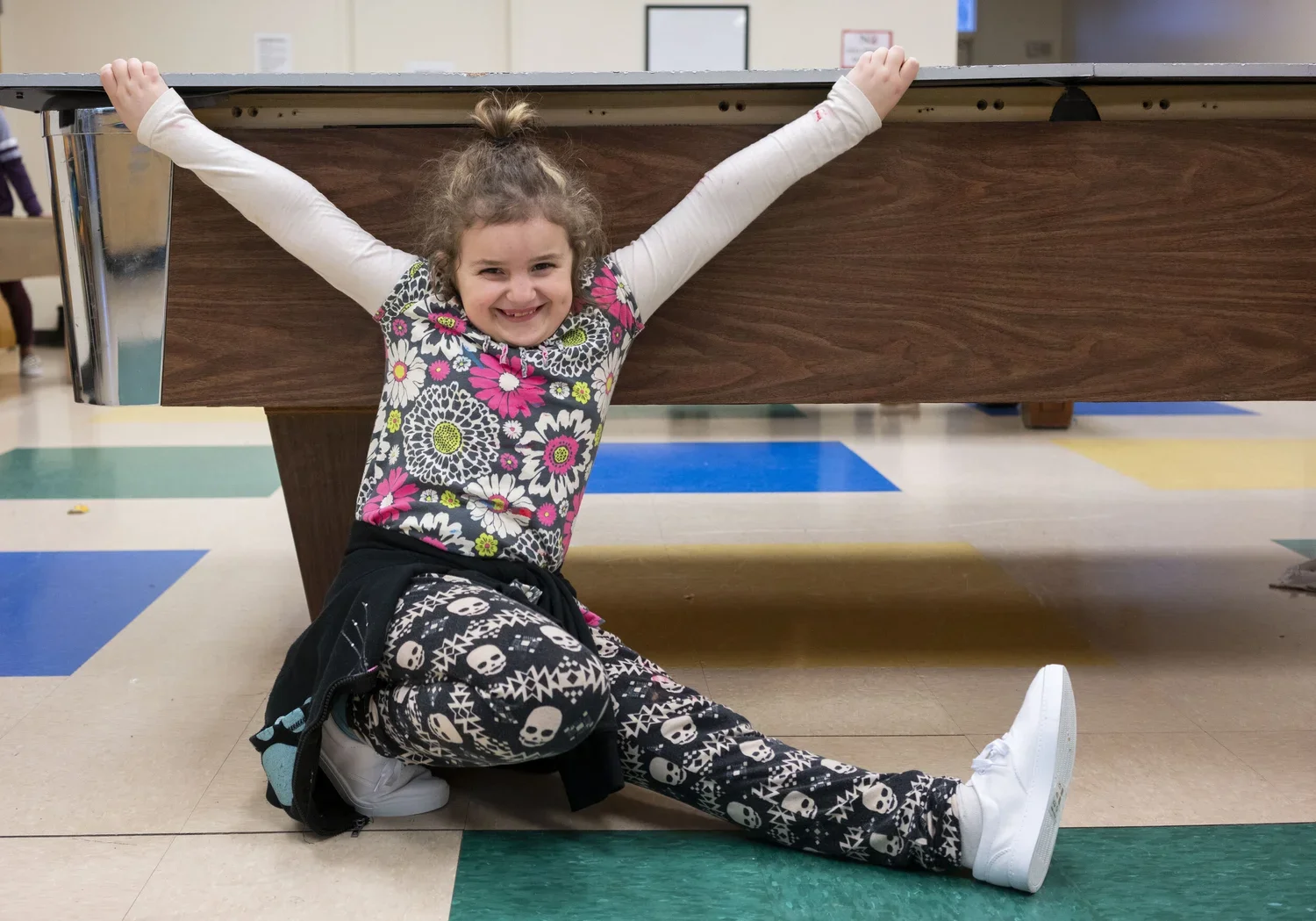 A young girl with curly hair smiling happily while sitting on the floor under a pool table, stretching her arms up along the sides of the table in a playful pose.