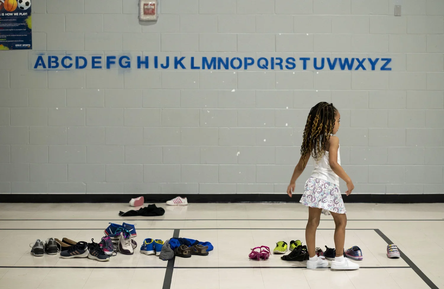 A young girl with dreadlocks stands inside a gymnasium, looking to her left. Behind her, there are shoes and socks laid out on the floor for an activity. The background features a light gray brick wall with the alphabet in blue letters.