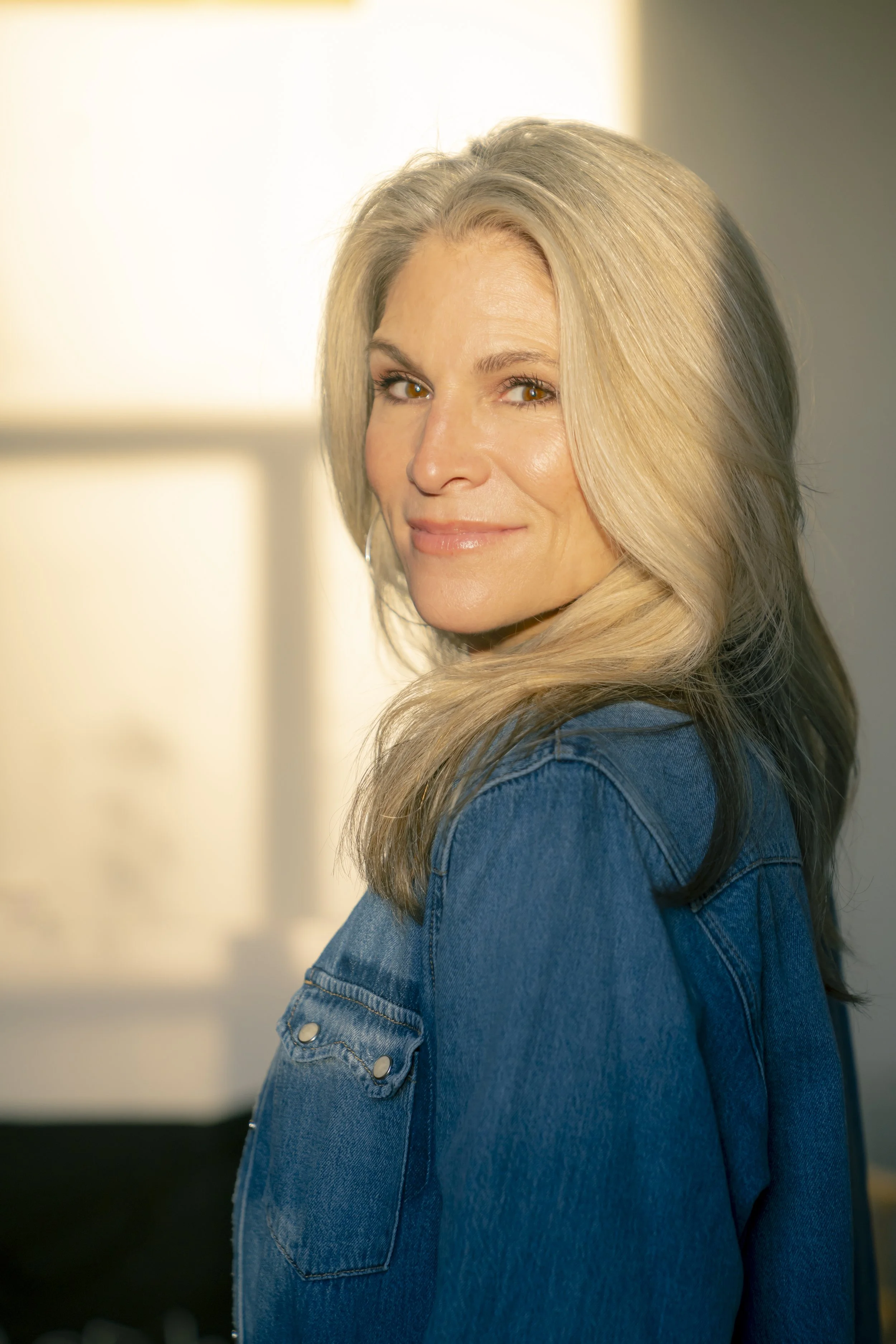 A woman with long, blonde hair wears a blue denim jacket and smiles softly, in a well-lit room.
