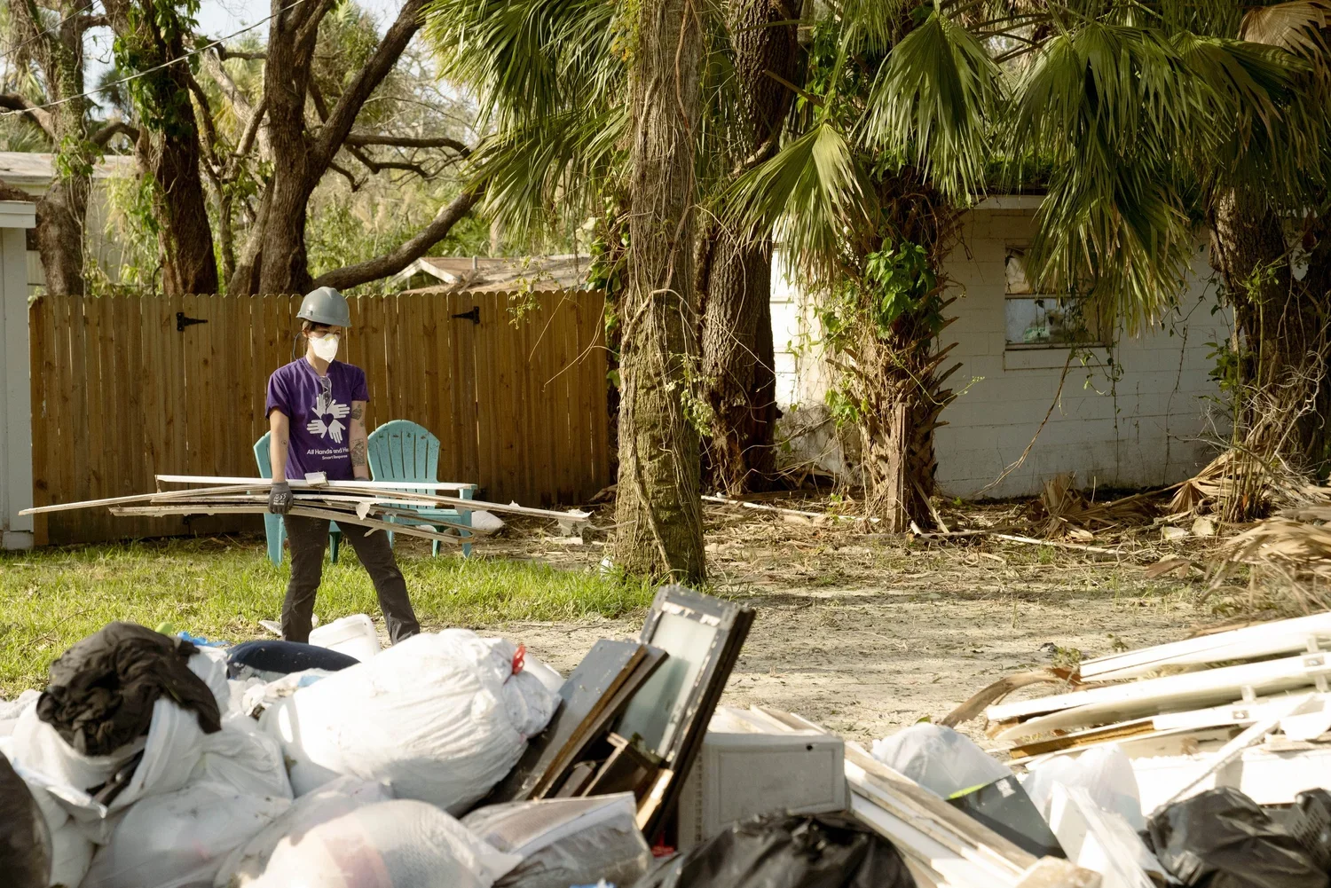 A person wearing a purple shirt, face mask, gloves, and a helmet carrying metal and wood debris in a yard, with debris piles in the foreground, trees, a wooden fence, and a small white building in the background.