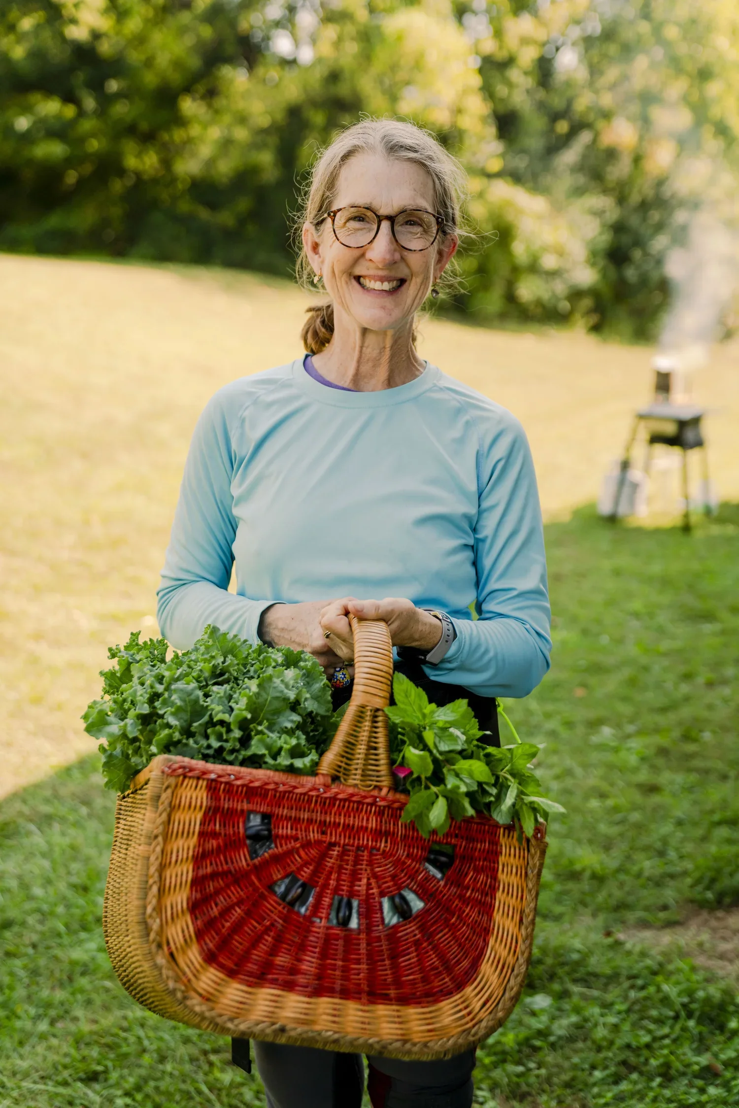 A smiling elderly woman with gray hair and glasses, wearing a light blue long-sleeve shirt, holding a basket filled with leafy greens, standing outdoors in a park with trees and a grill in the background.