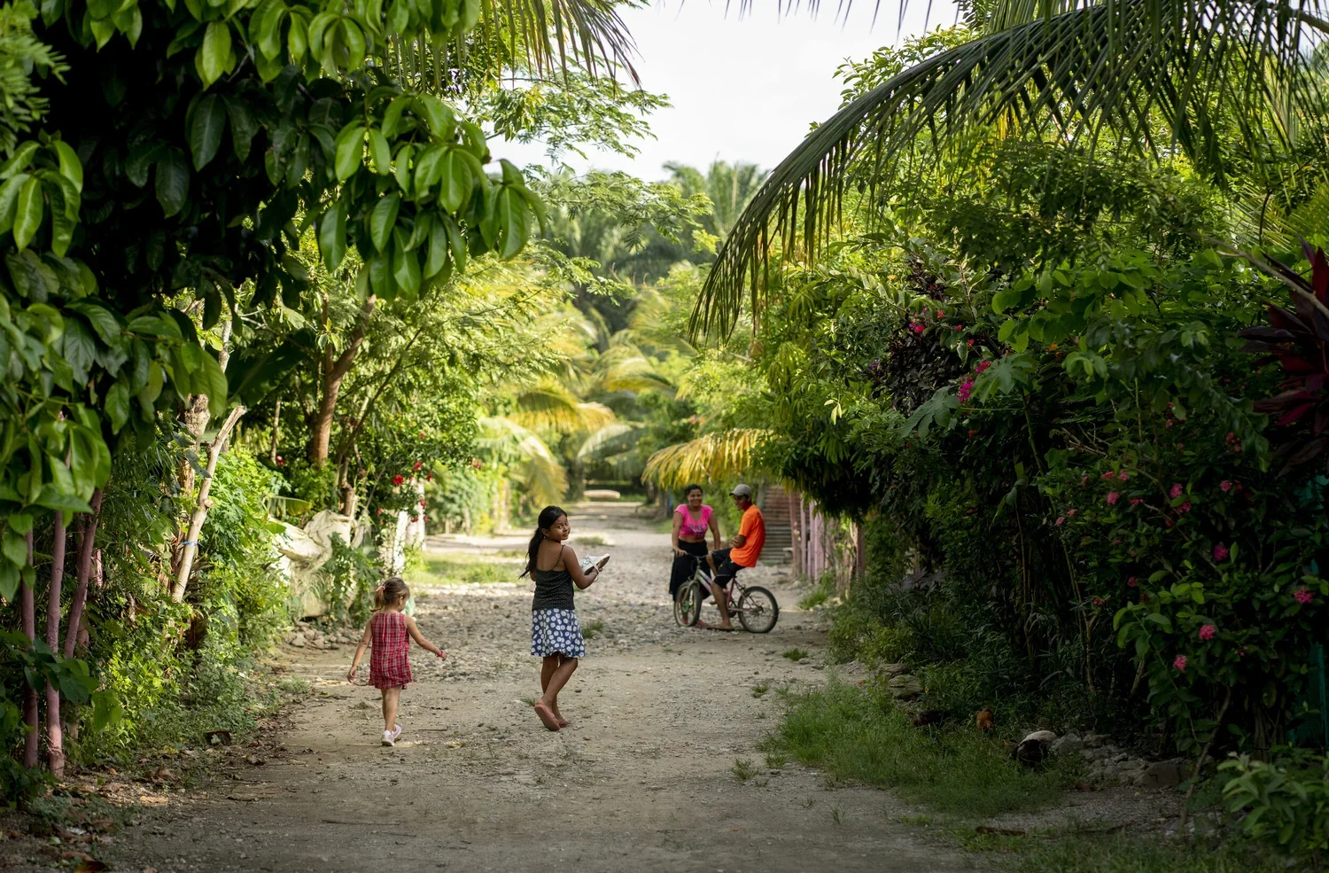 Children and adults walking and chatting on a dirt path surrounded by lush green tropical plants and trees.