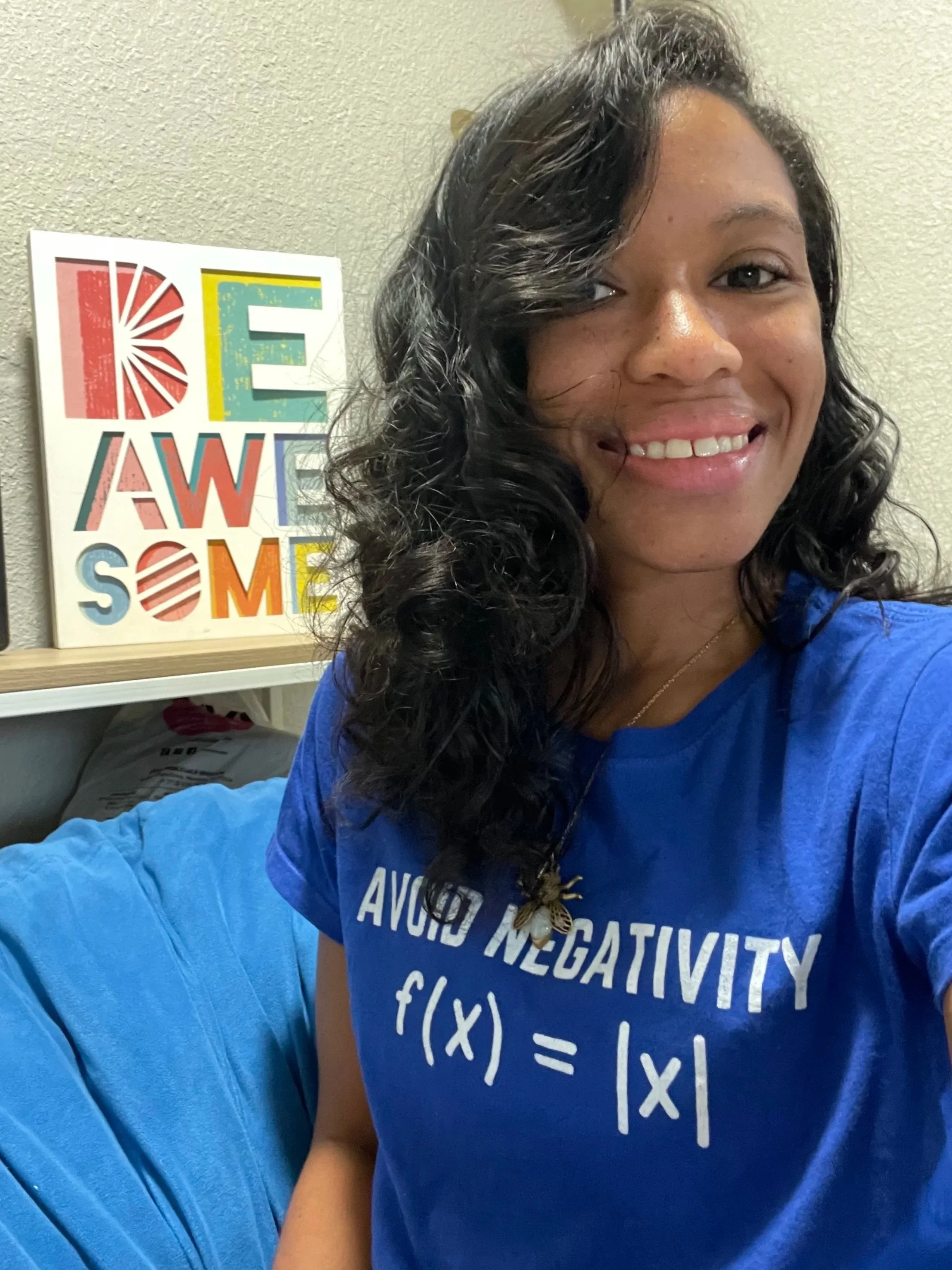 Woman with dark, curly hair smiling, wearing a blue t-shirt with the phrase "Avoid Negativity" and the notation for the absolute value parent function, sitting on a beanbag next to a colorful sign that reads 'BE AWESOME'.