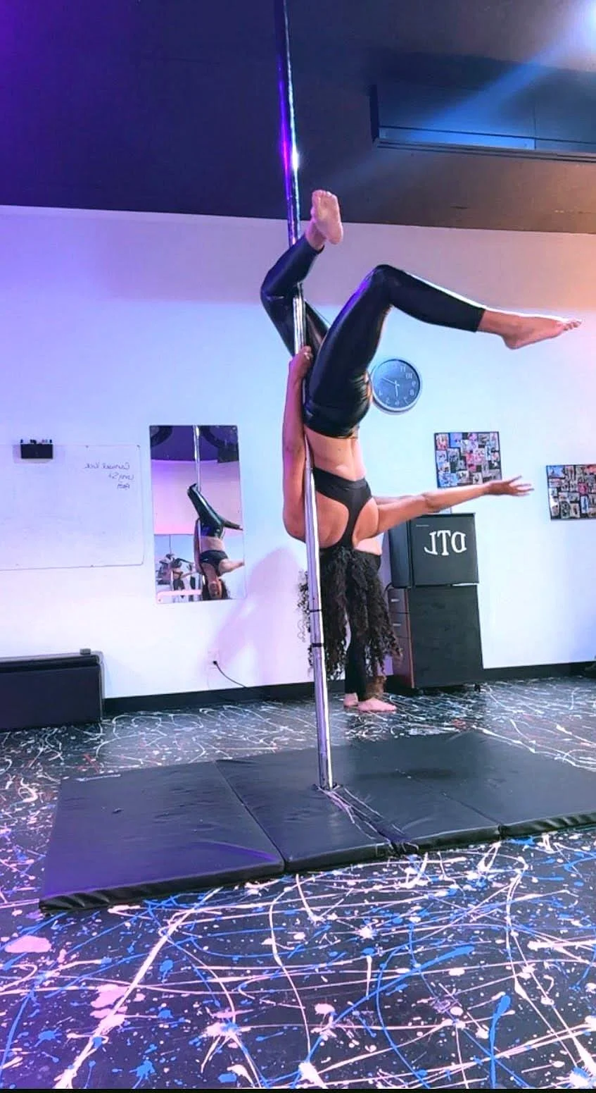 A woman performing a pole dance move on a pole, with her head and arms on the floor and her legs extended upward. She is wearing black athletic wear and is in a studio with colorful paint splatter flooring, a mirror, and some wall decorations.