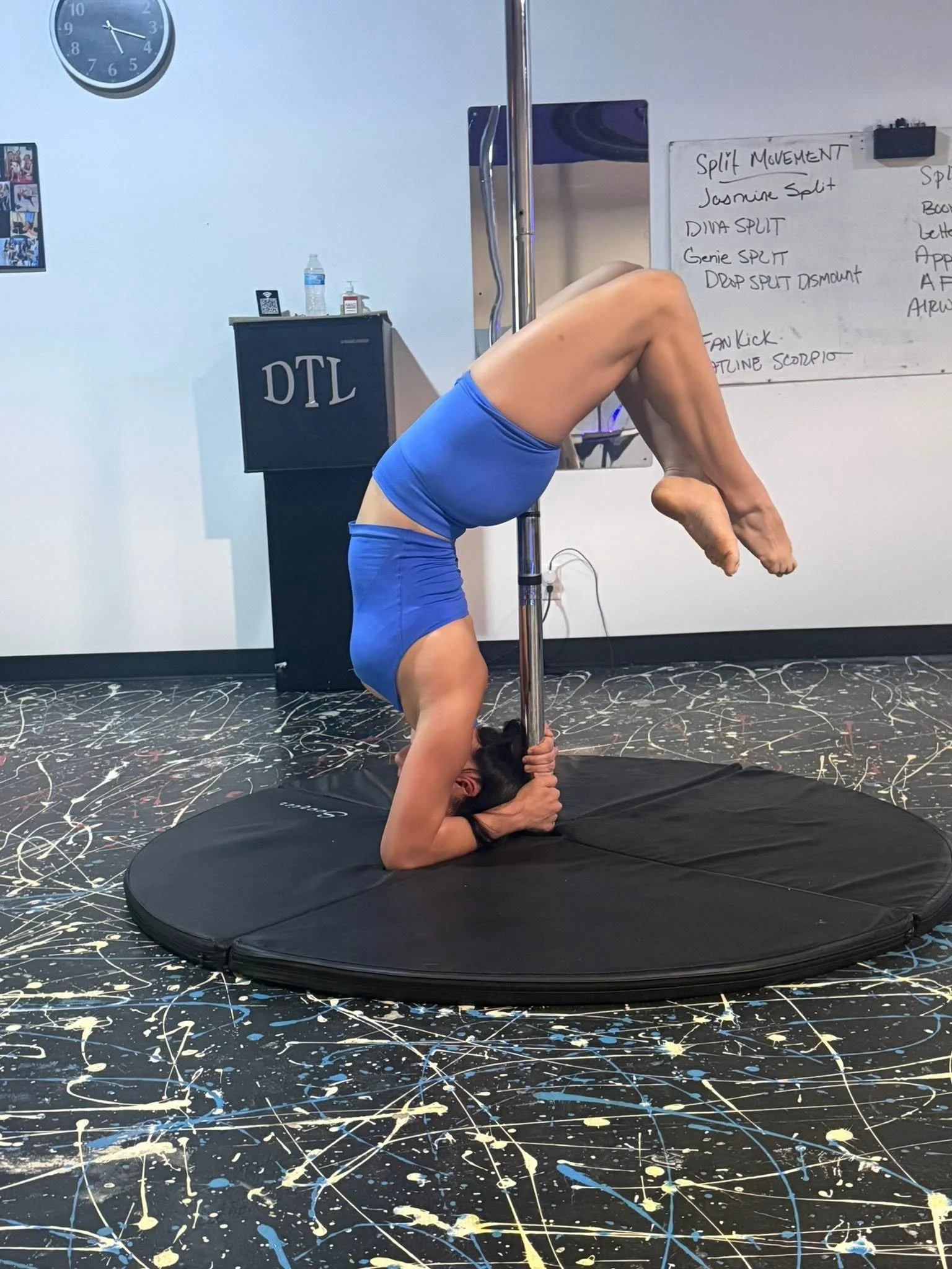 A woman practicing pole dance in a studio, doing a pose upside down on the pole with her head and hands on the floor, wearing blue athletic wear, on a black mat, with a whiteboard, clock, and other equipment in the background.