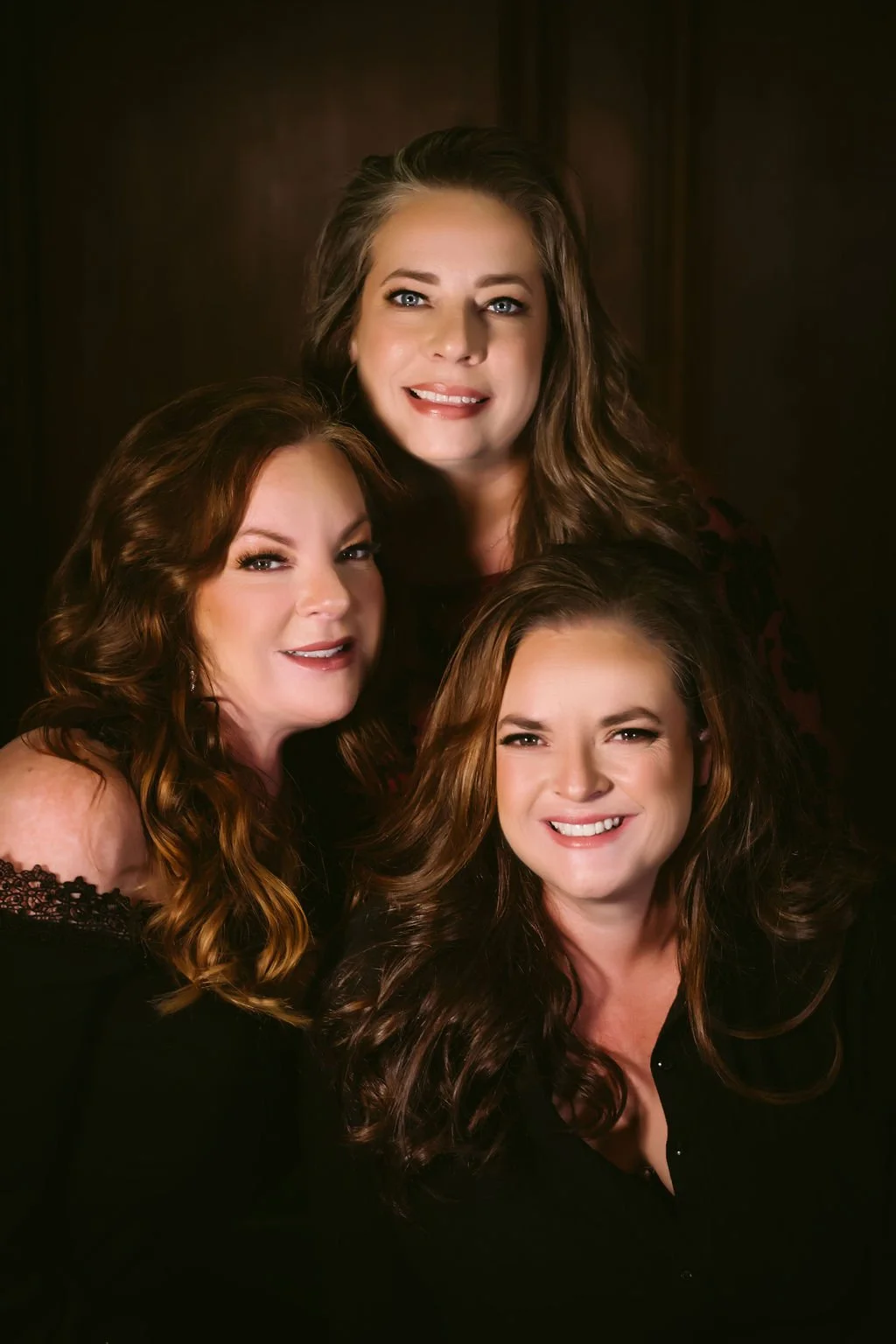 Group portrait of three women with long curly hair, smiling, against a dark background.